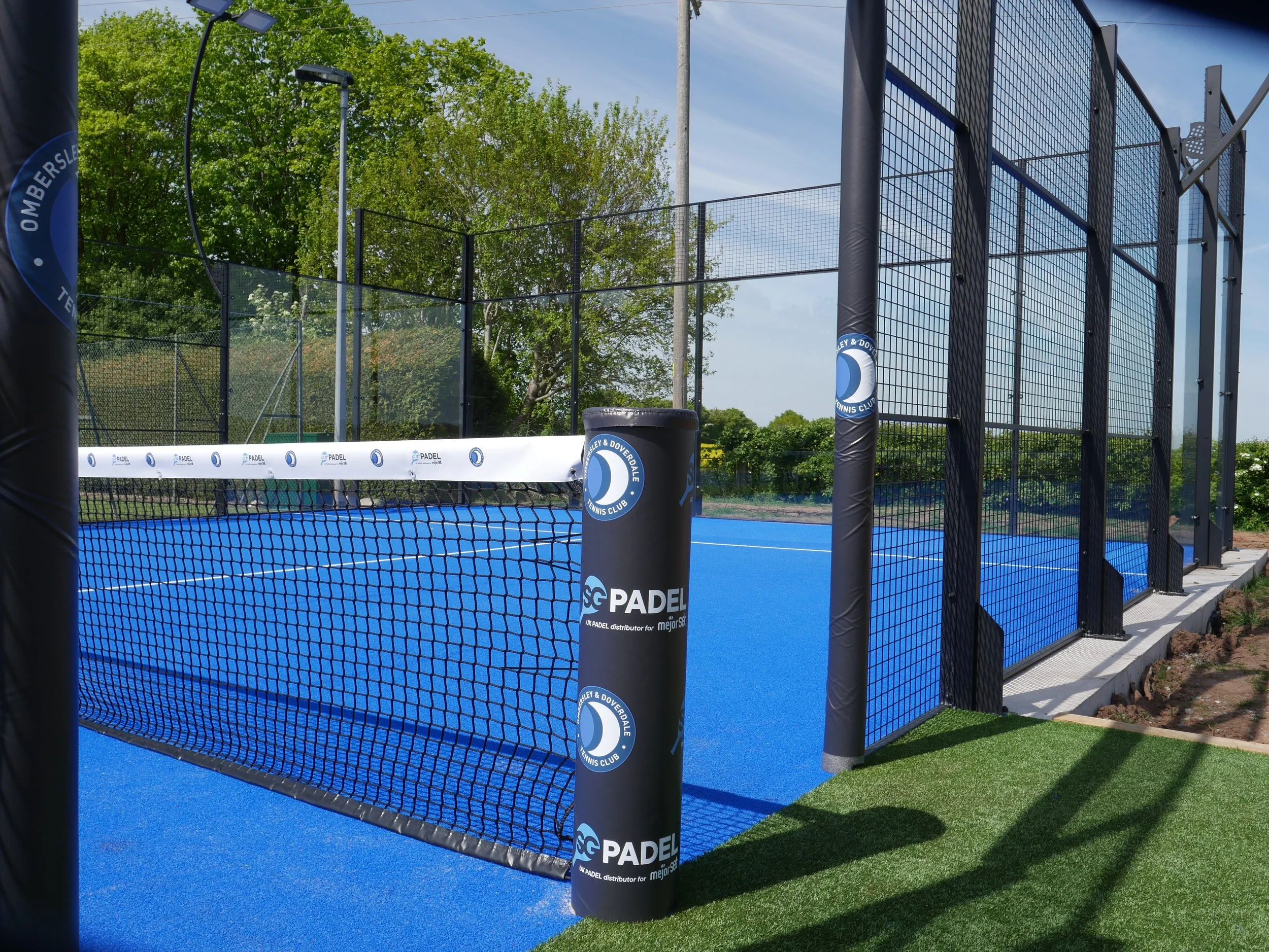 An outdoor padel tennis court with blue playing surface, surrounded by tall black fencing, and a net in the middle. The court is situated in a green area with trees in the background, and there is a shadow of the net post on the ground.