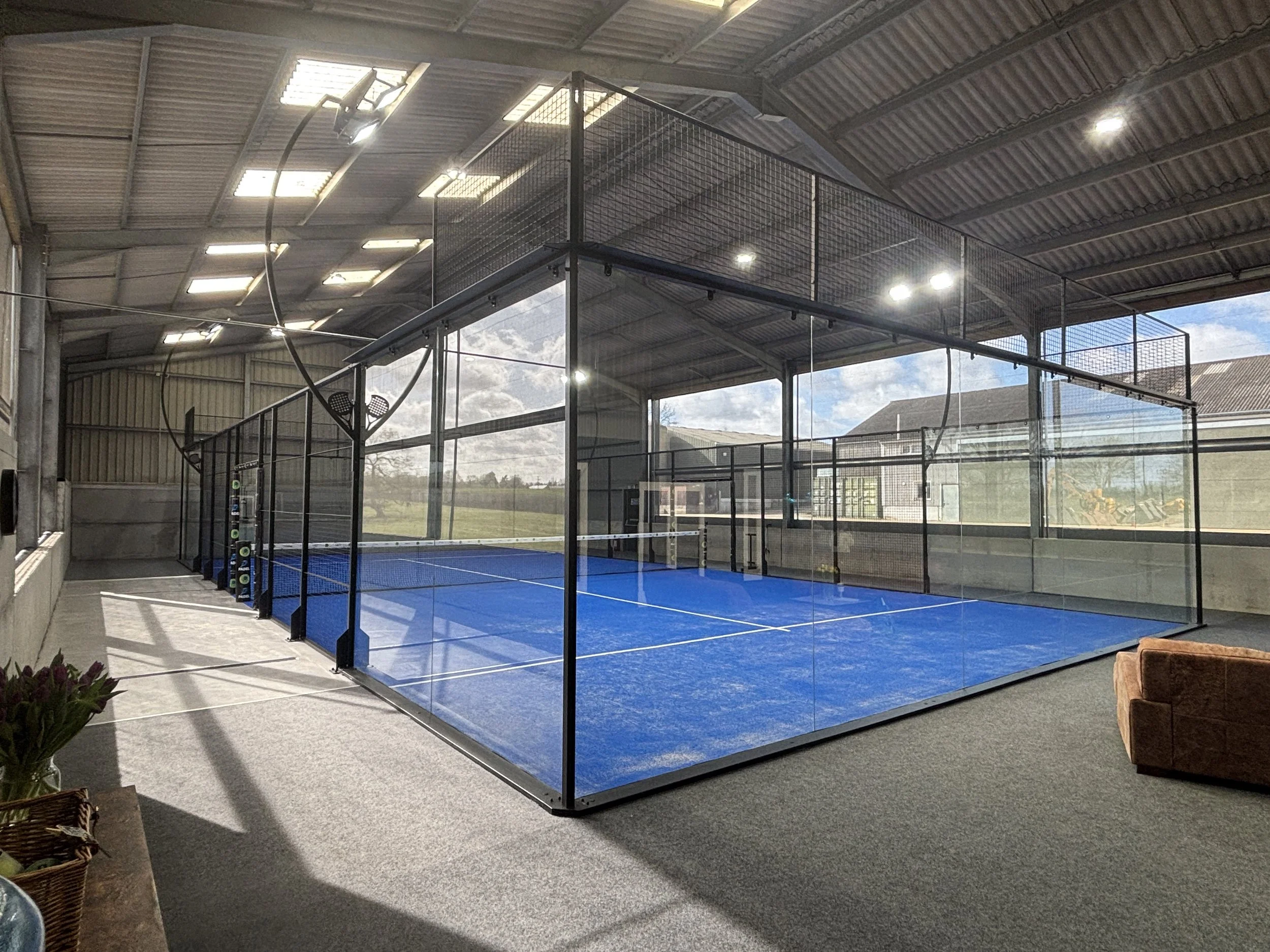 Indoor Padel court with blue surface, enclosed with glass walls, illuminated by overhead lights, with an outside view of a cloudy sky and part of a building roof.