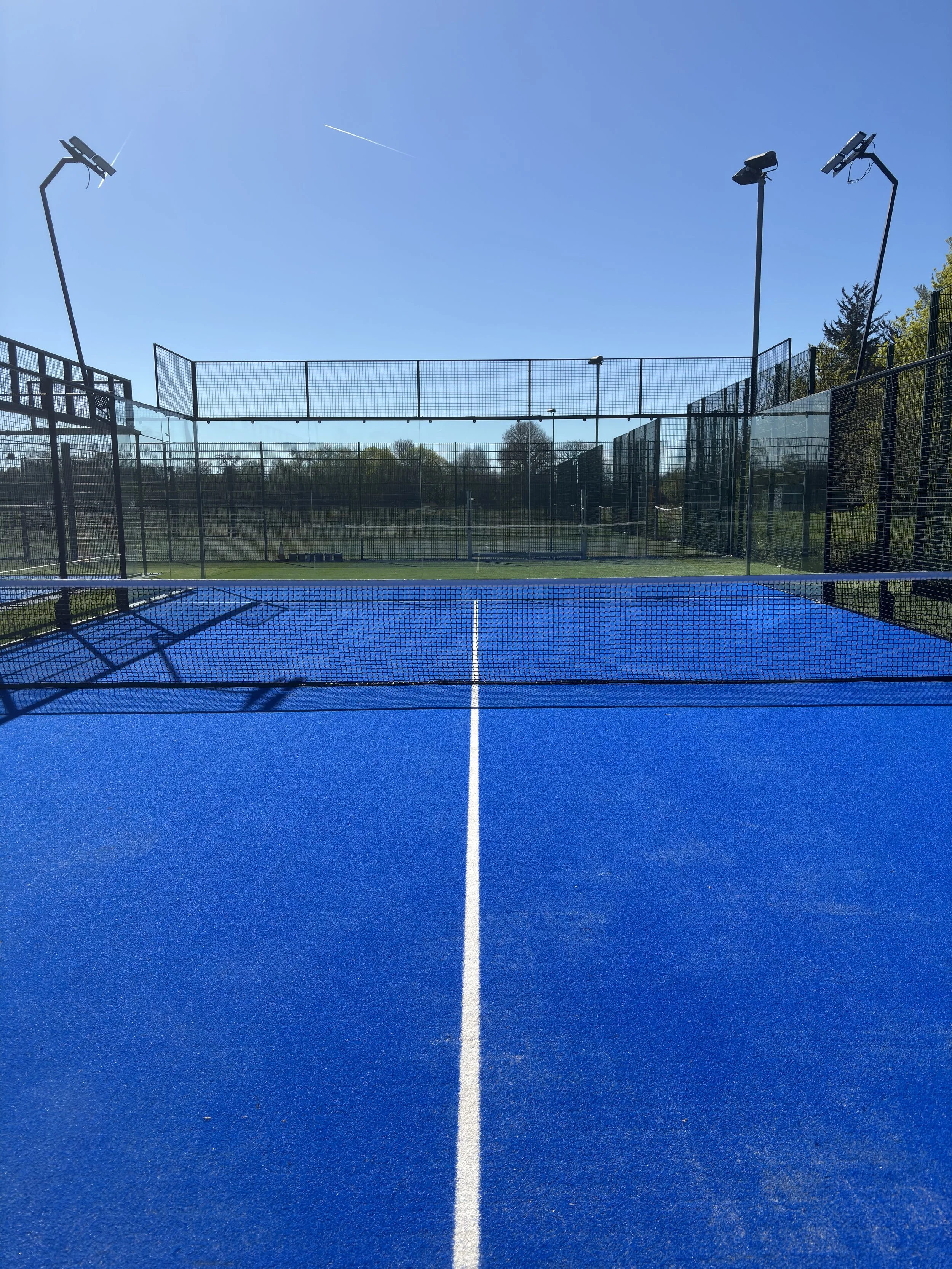 View of a blue outdoor Padel court with a white court line, surrounded by black fencing, and four tall light posts under a clear blue sky.