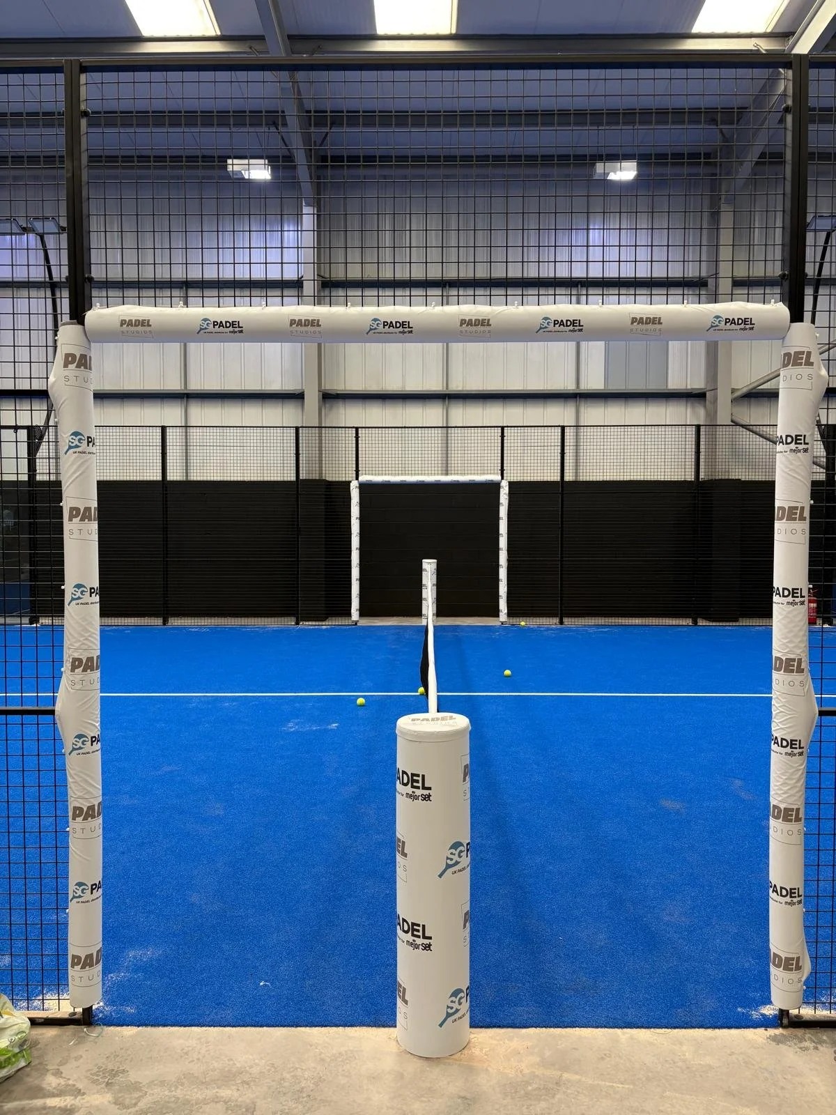 Indoor padel court with blue flooring, black fencing, and white goal posts Padel Studios Coventry