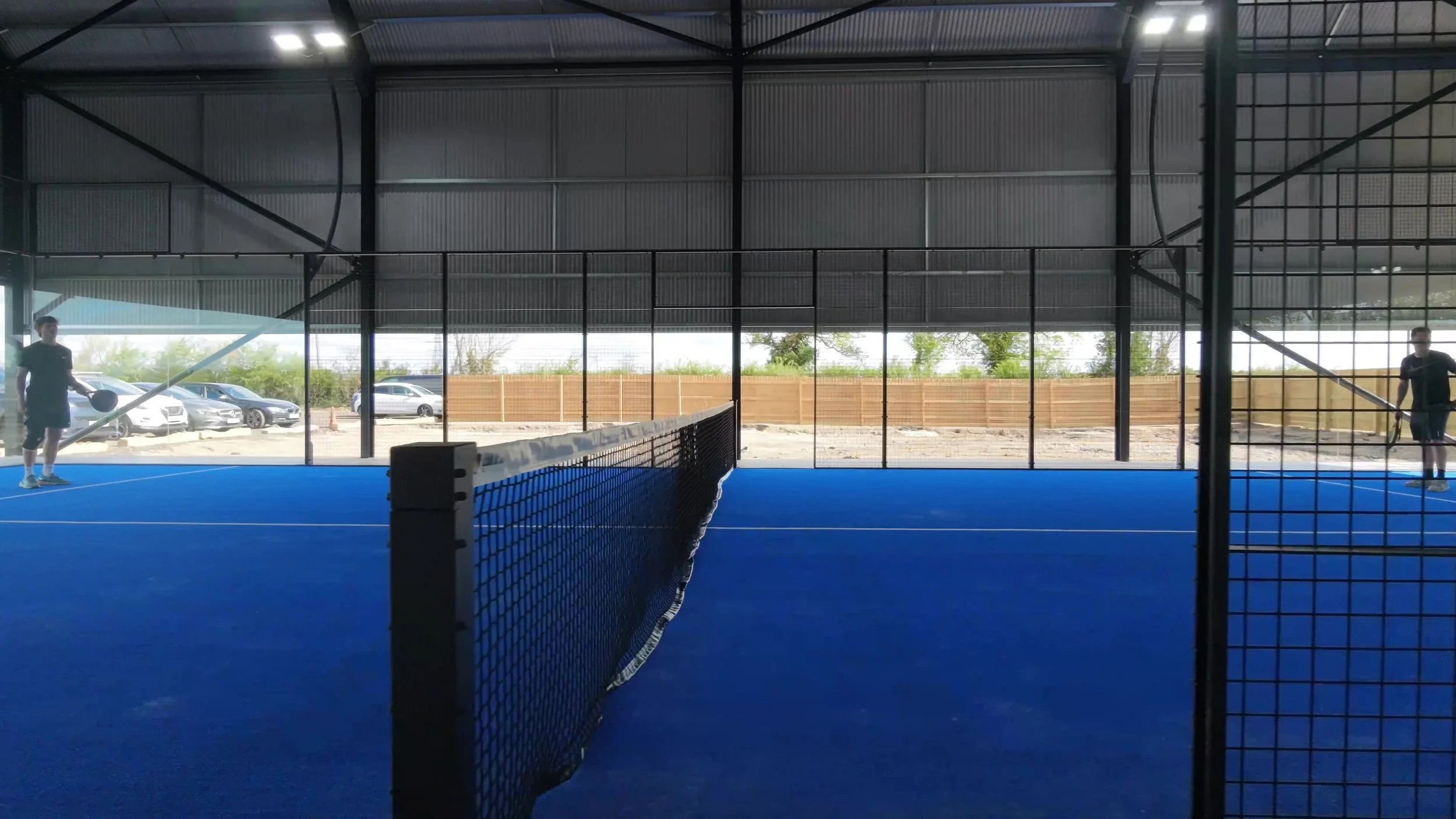 Indoor Padel court with blue surface and net, two players practicing, large windows showing parked cars outside, and a metal roof overhead.