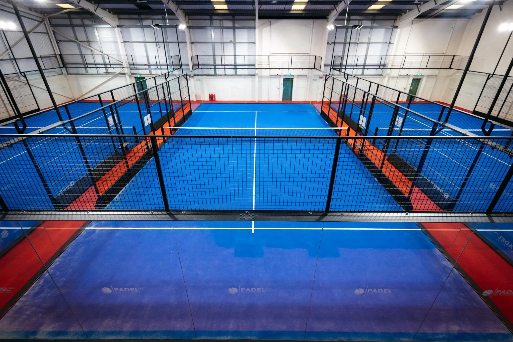 Indoor padel tennis court with blue playing surface surrounded by glass walls and black fencing UKs Best Padel