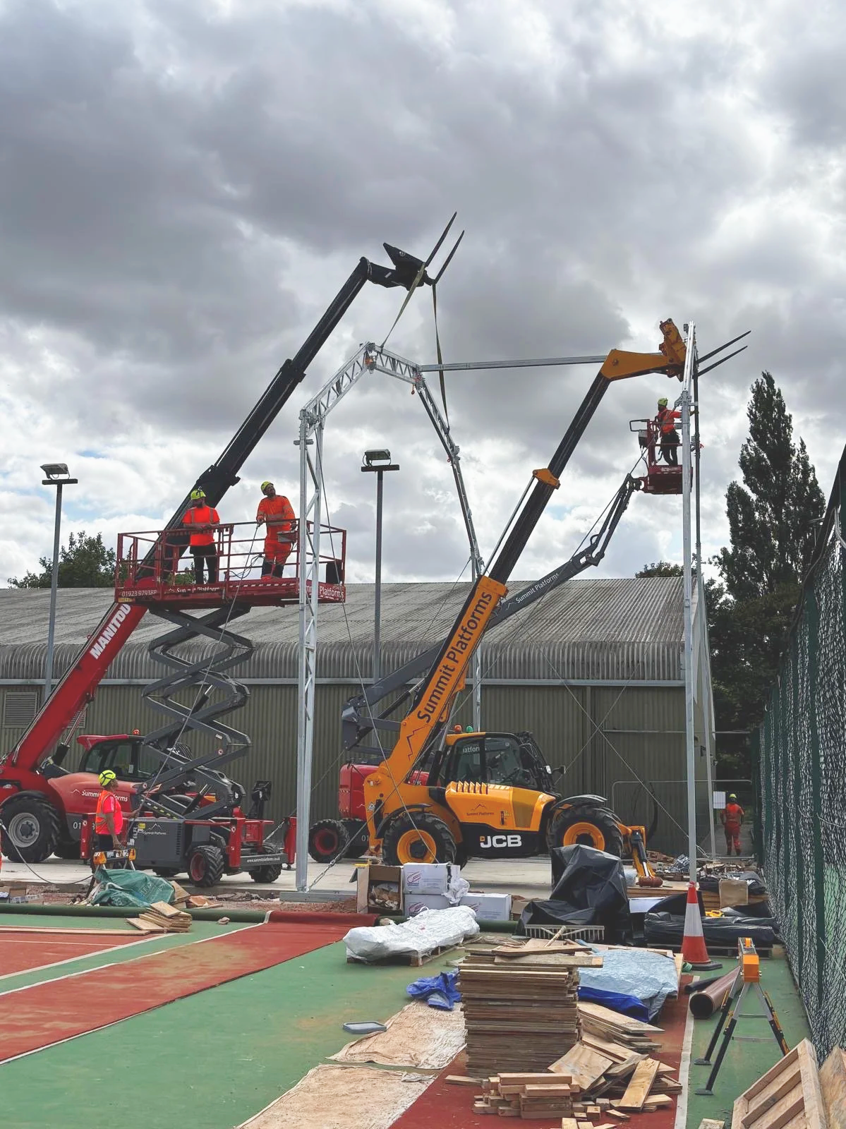 Construction workers using cranes and lifts to install and assemble a large Canopy For an outdoor Padel Site, with building materials and tools scattered nearby.