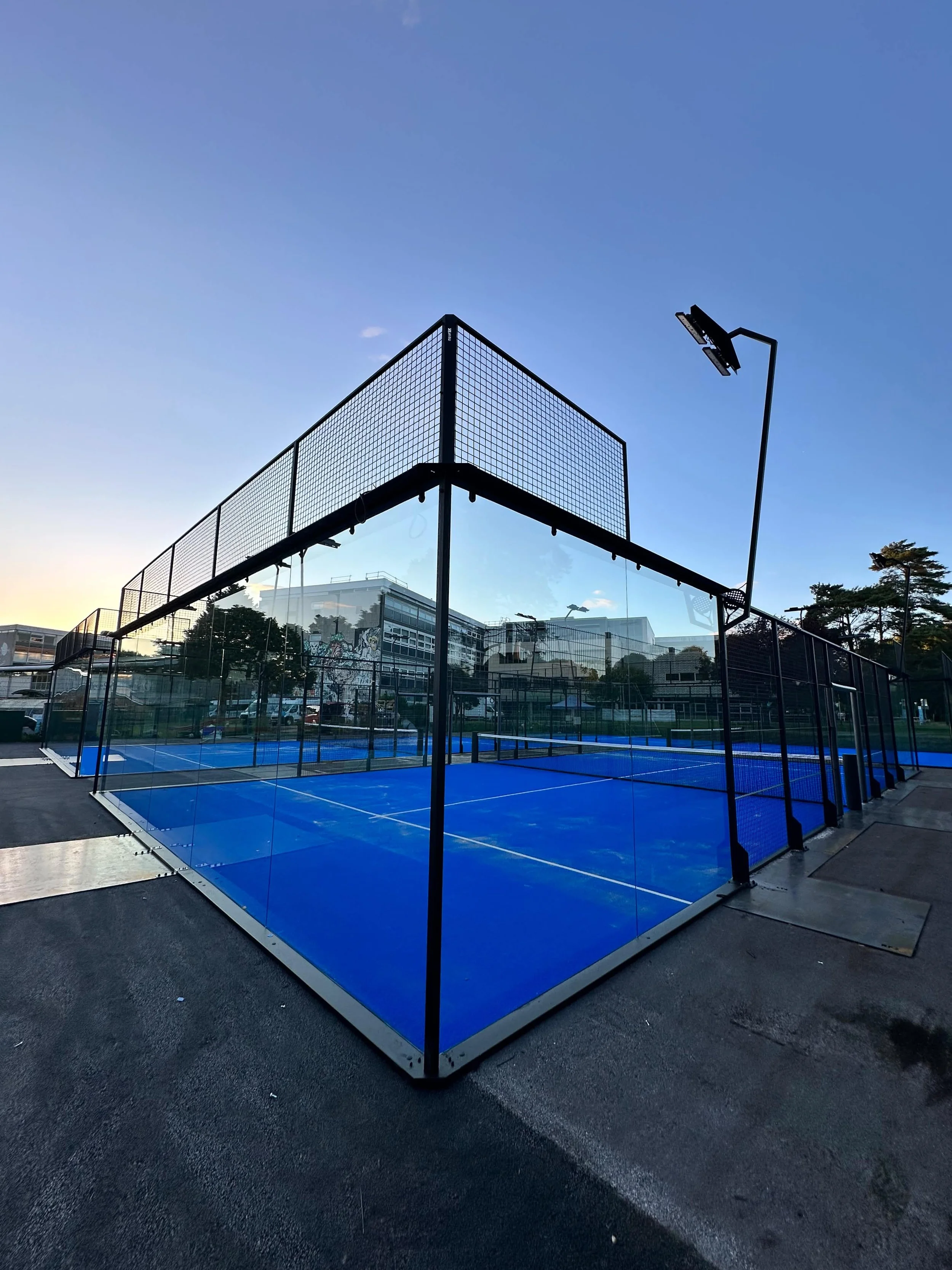 An outdoor tennis court with a bright blue surface, surrounded by a glass fence. There is a tall floodlight on a pole and a climbing wall in the background, with trees and buildings under a clear evening sky.