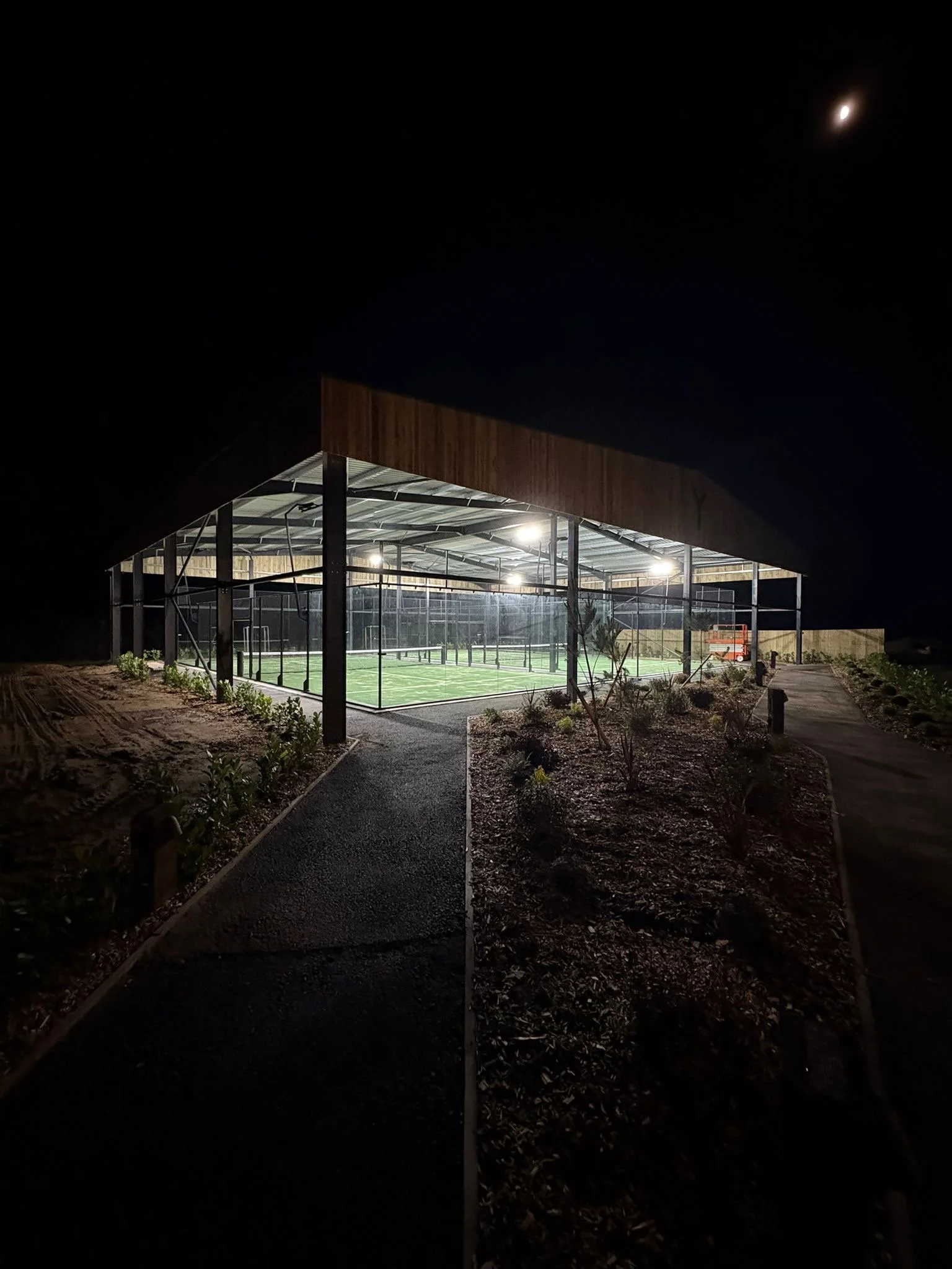Night view of an illuminated padel tennis court at Lake Y with a partially covered roof, surrounded by landscaped pathways and garden beds.