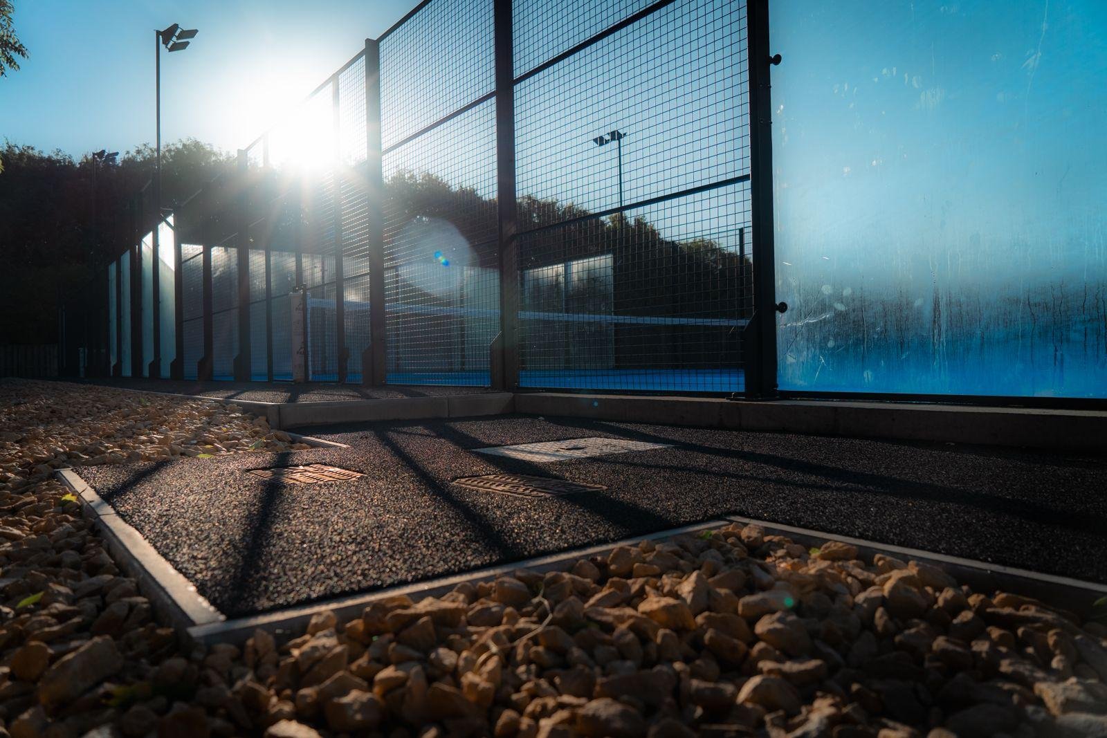 An outdoor Padel court surrounded by a tall fence, with the sun shining brightly in the sky creating lens flare, and textured ground surface visible in the foreground. UKs Best Padel courts