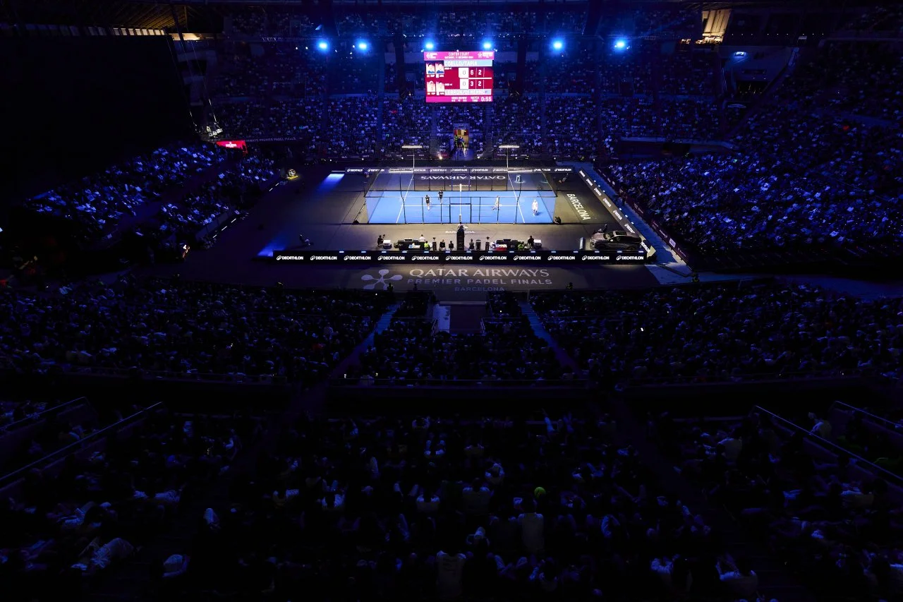 Indoor Padel Tennis arena filled with a large audience, illuminated with blue lighting, with a Blue Padel court in the center. A scoreboard displays scores and game information above the court.