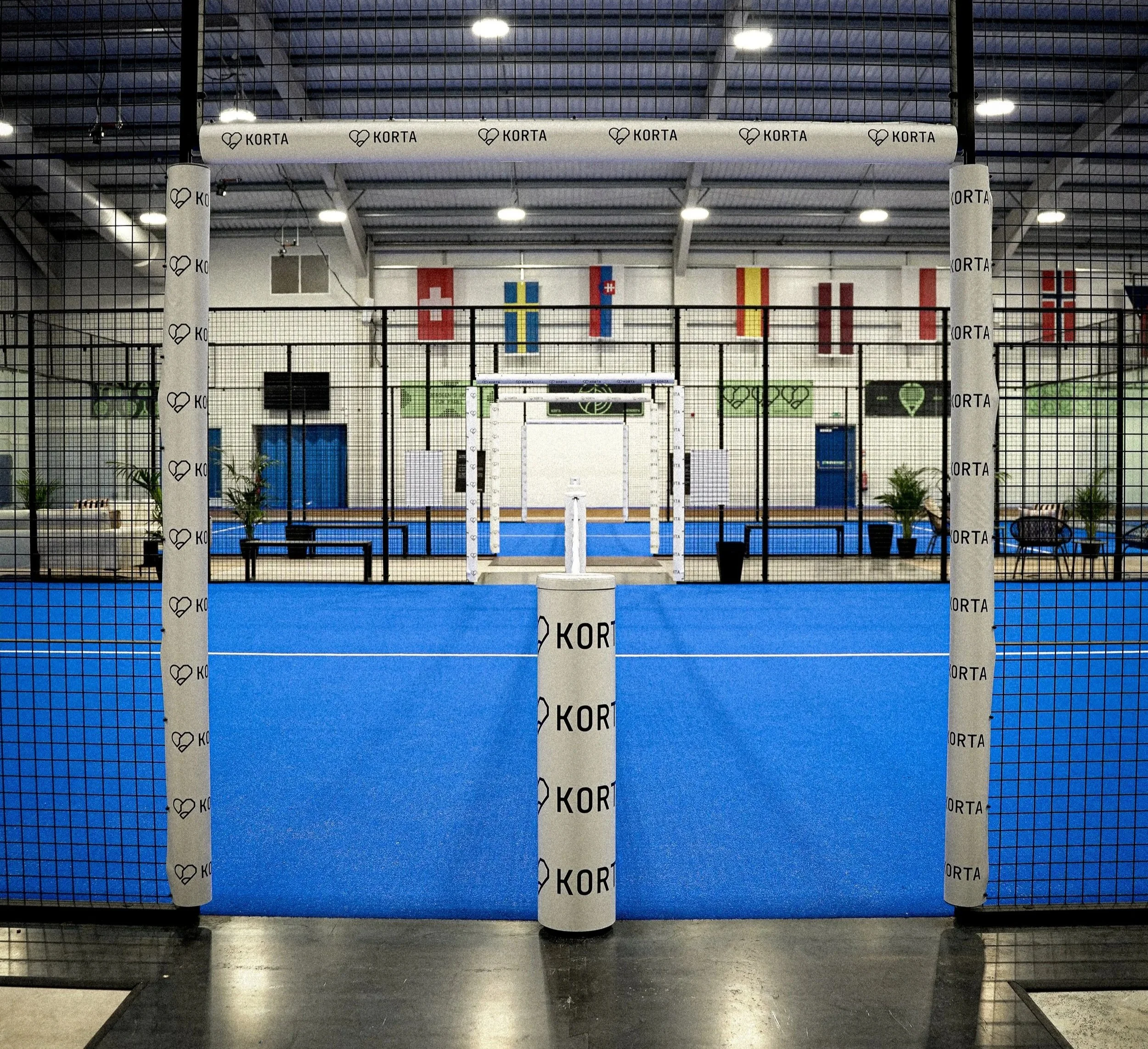 An indoor Padel facility with a blue court, black fencing, and tall padded poles with the brand name 'KORTA' printed on them. Six European flags are hanging on the back wall, which is white with some windows and a vent.