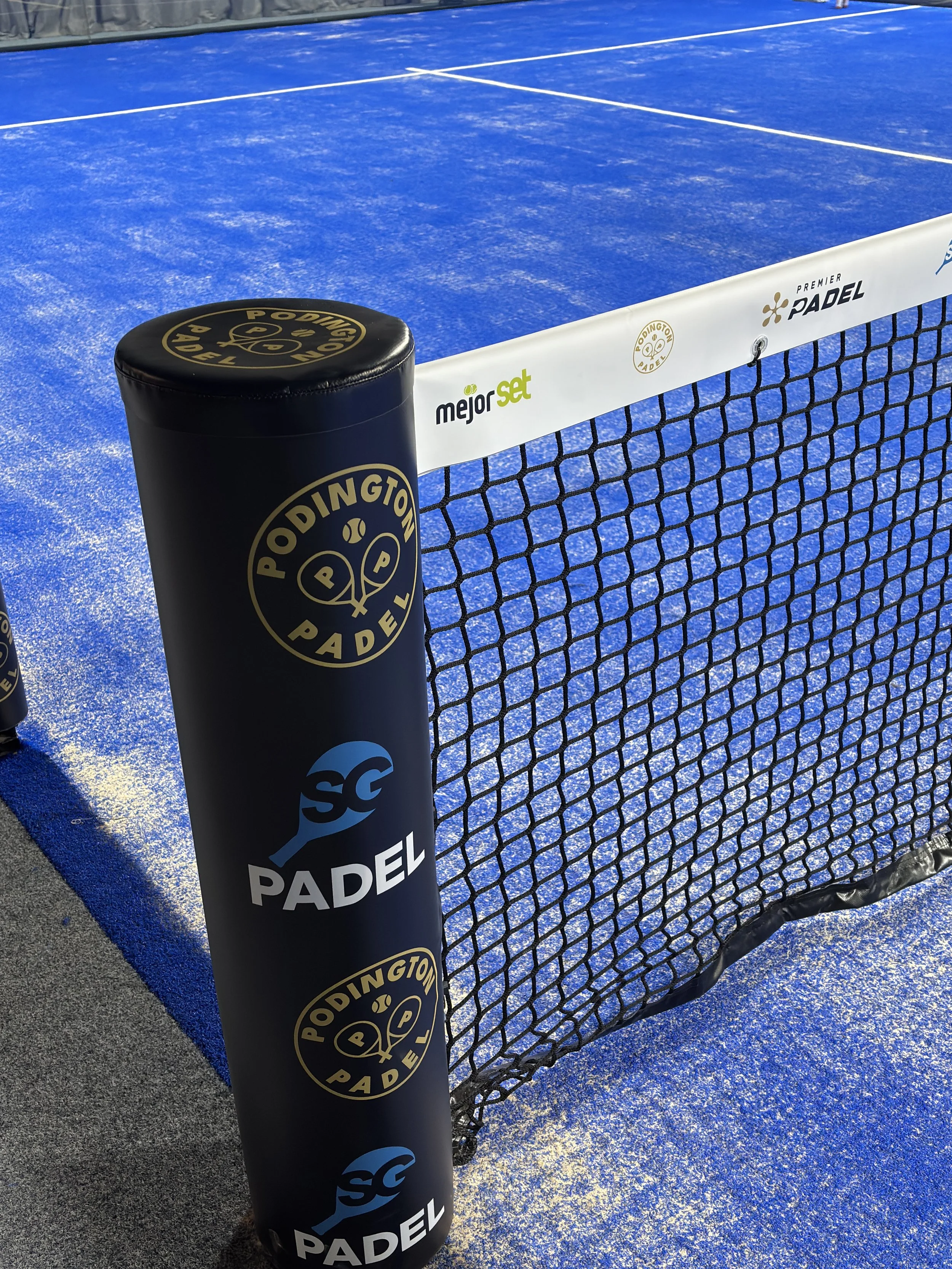 Close-up of a padel courts Personalized net and post with logos, Podington Padel and SG Padel on a blue court surface.