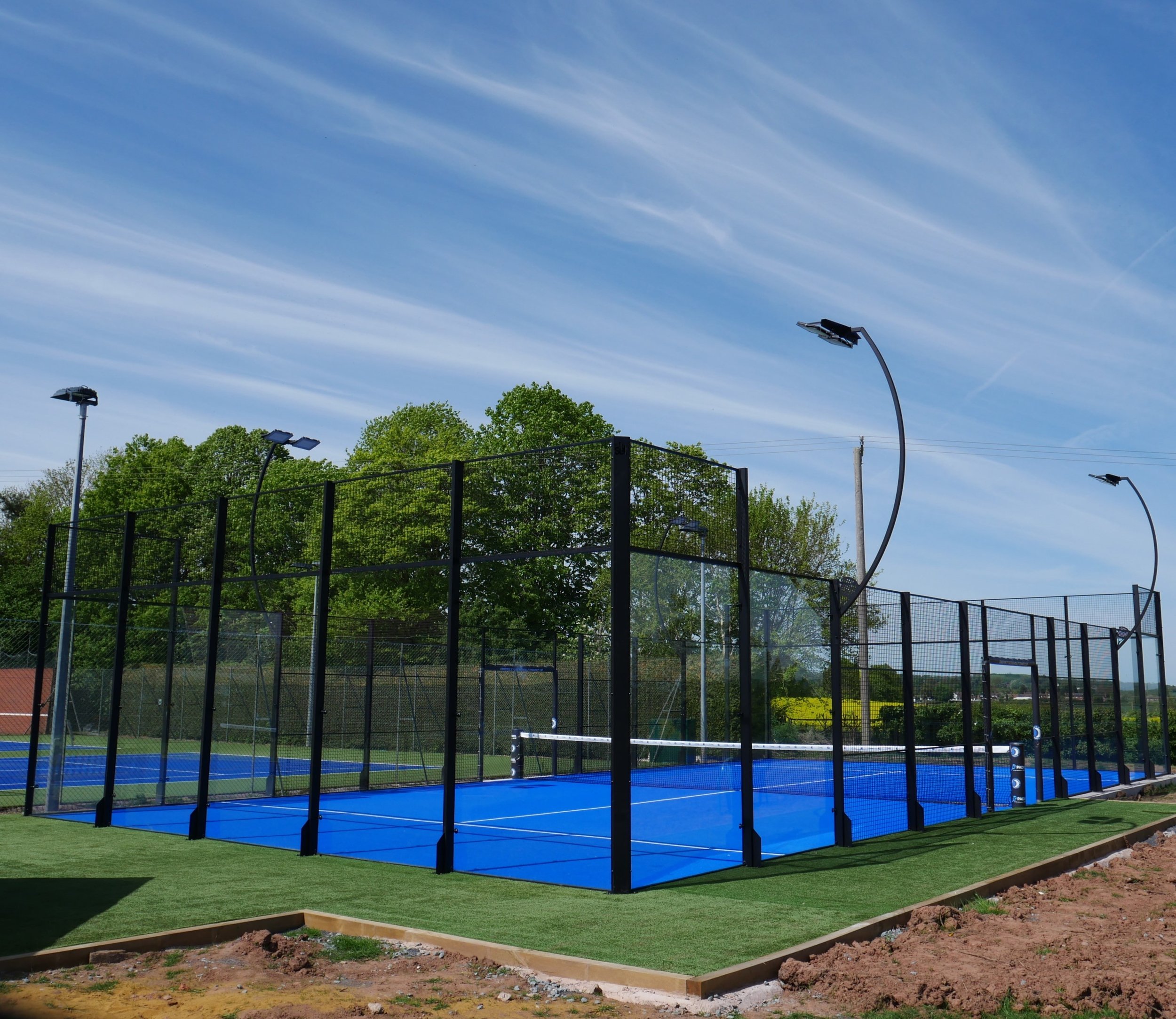Outdoor padel tennis courts with blue surface, surrounded by black fencing, tall floodlights, green trees, and a blue sky.
