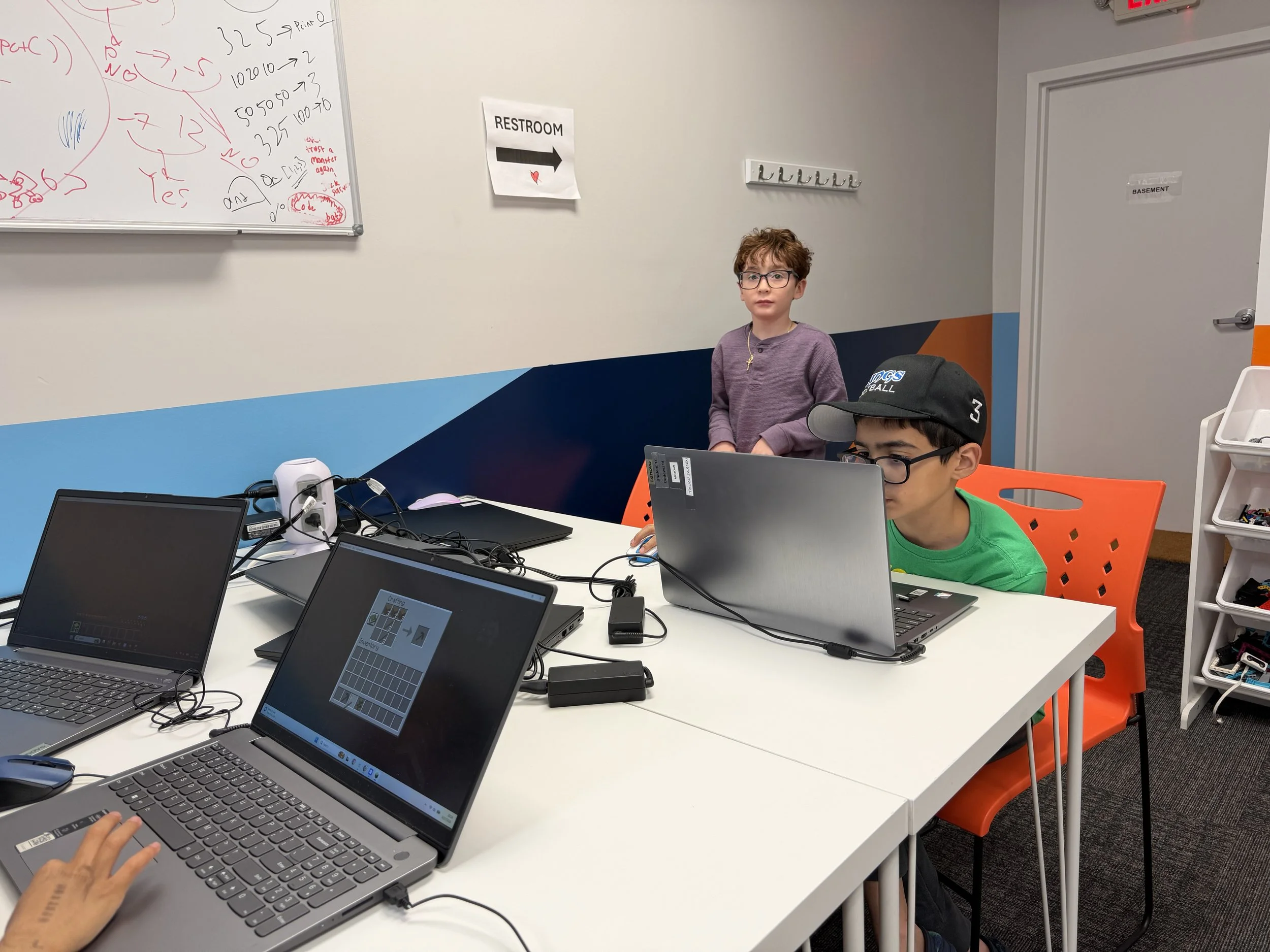 Two children working on laptops in a classroom or study space, with a whiteboard and a sign indicating a restroom in the background.