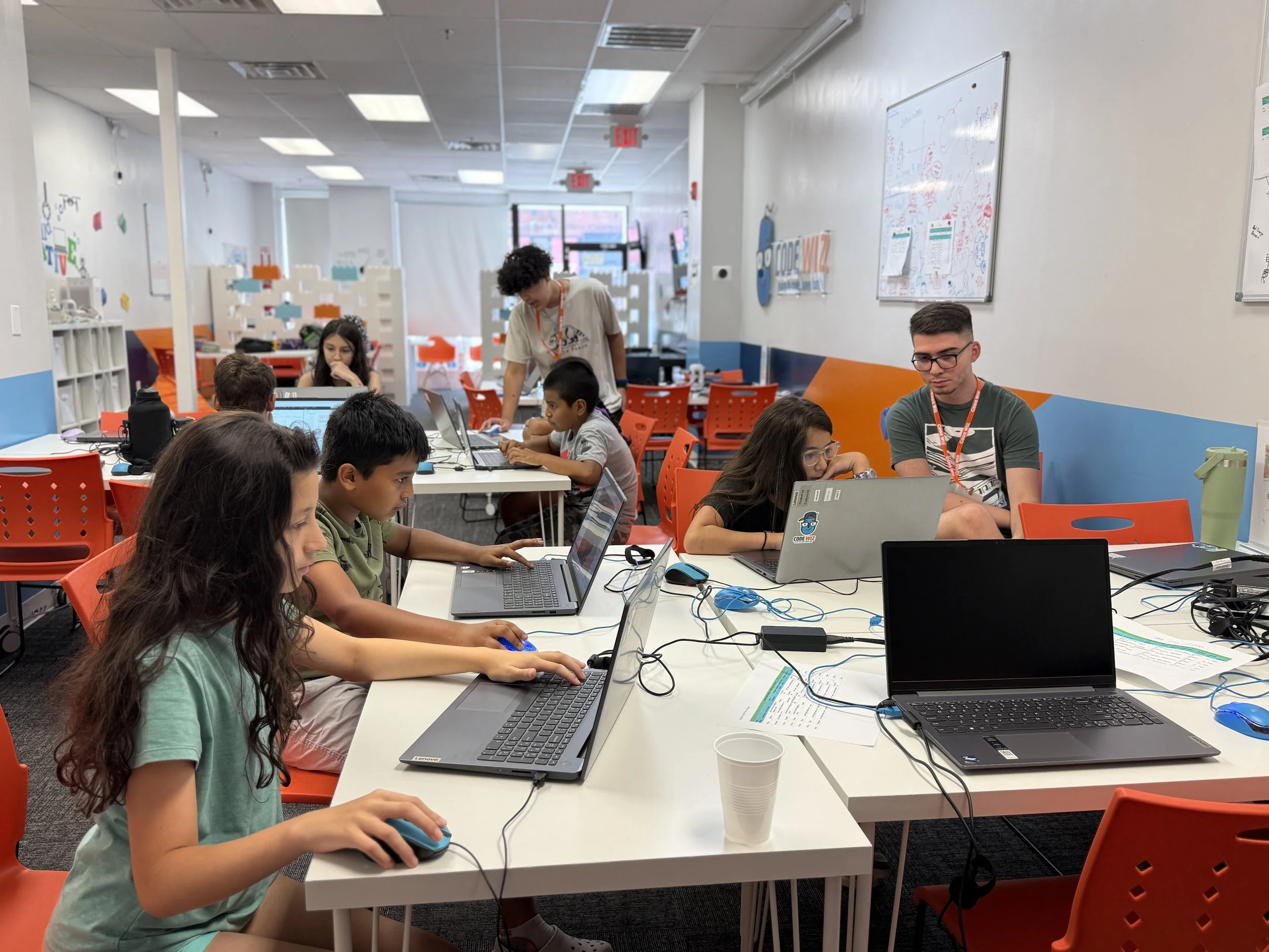 Children working on laptops in a classroom with a teacher guiding at the back, whiteboards on the walls, orange chairs, and a bright, colorful environment.
