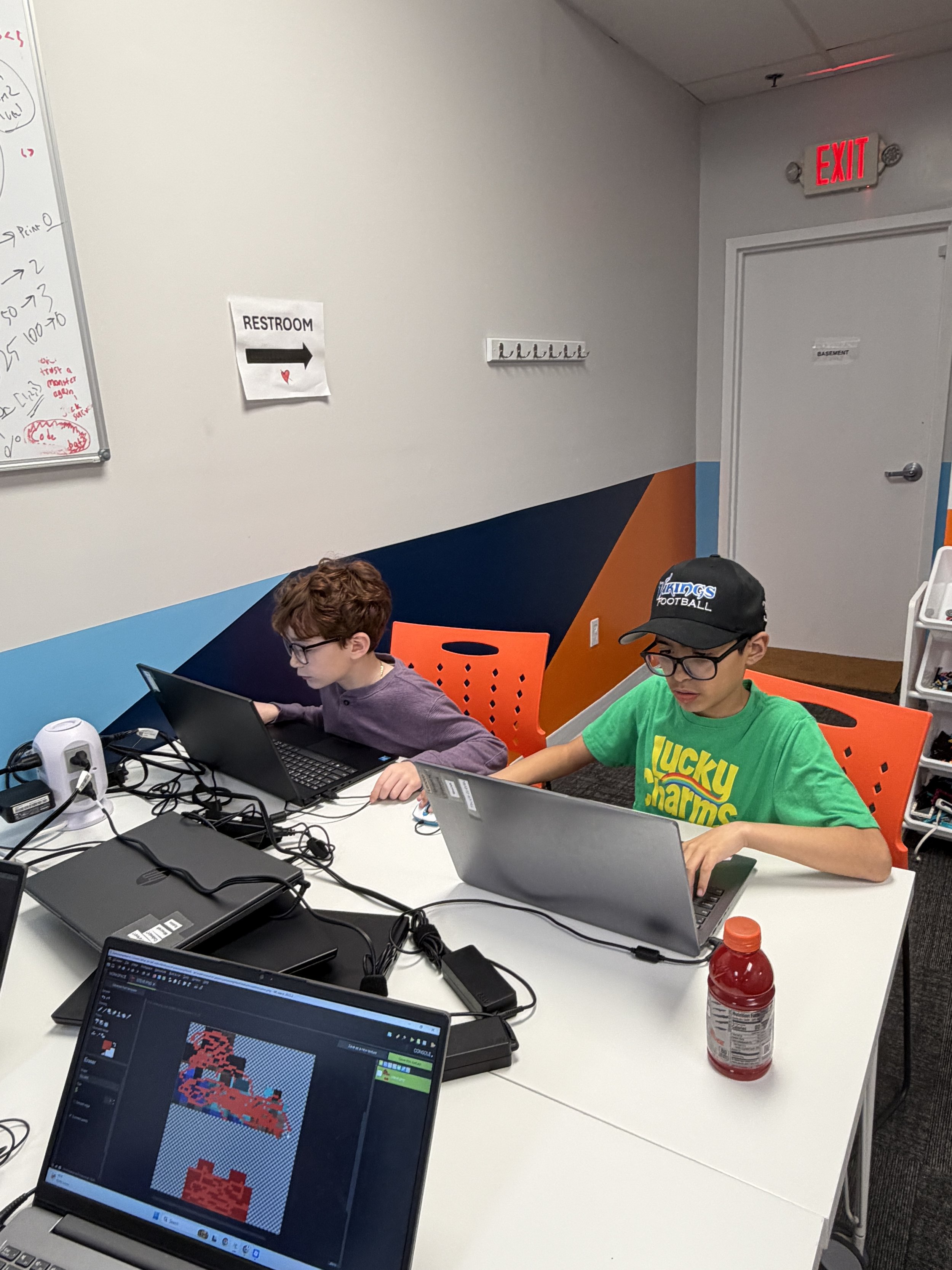 Two boys working on laptops at a table with computers, cords, and a red bottle. One boy wears a green T-shirt and a black cap, the other wears glasses and a purple sweater.