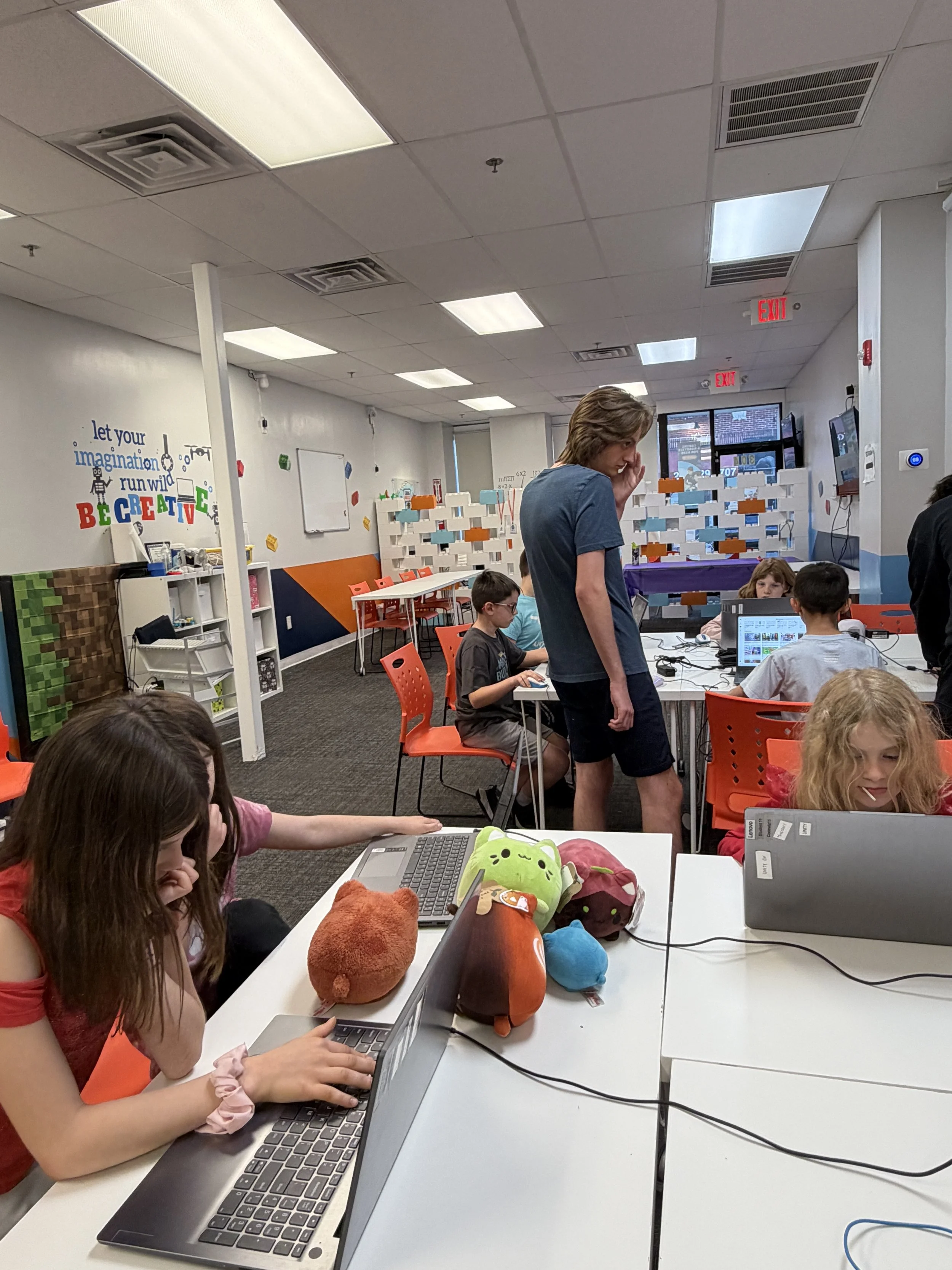 Children working on laptops in a classroom with colorful wall decor and stuffed animals on the desk.