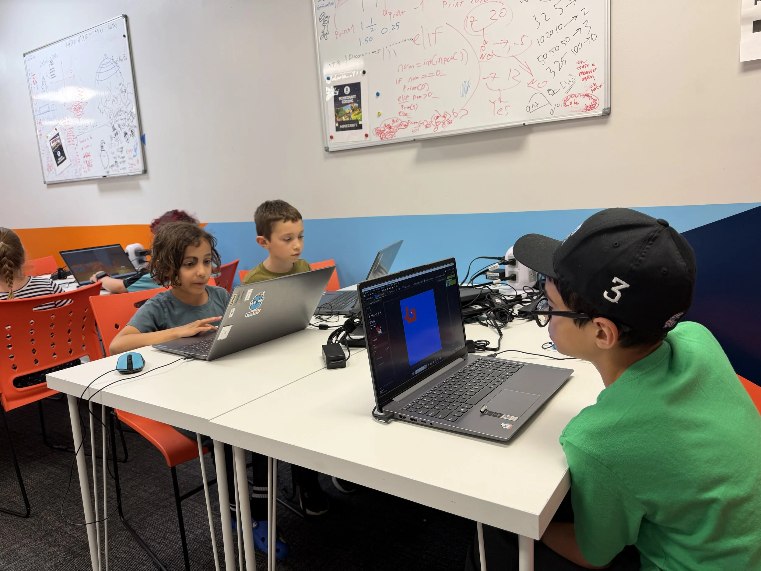 Children sitting at a table with laptops in a classroom, working on a coding project, with whiteboards filled with notes and drawings on the wall.