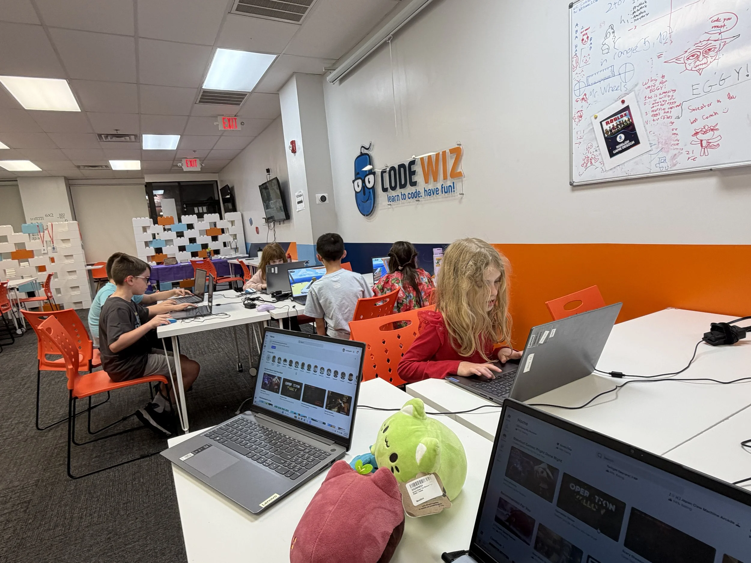Children working on laptops in a classroom with a sign that says 'Code WIZ' and a whiteboard with doodles and notes.