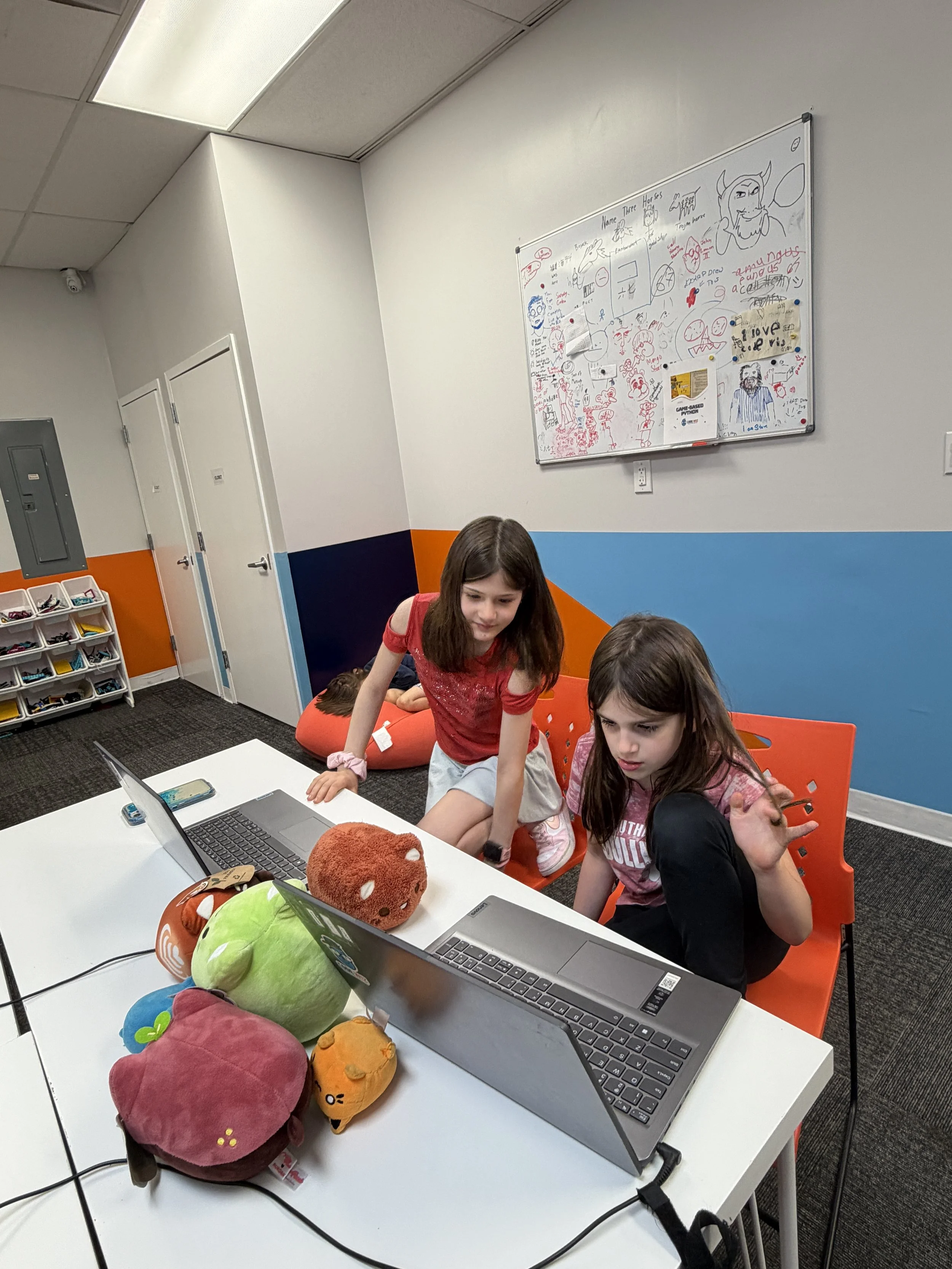 Two young girls are sitting at a white table looking at a laptop, with plush toys on the table and a third girl sitting on the floor behind them. A whiteboard with drawings and writings is on the wall behind them.