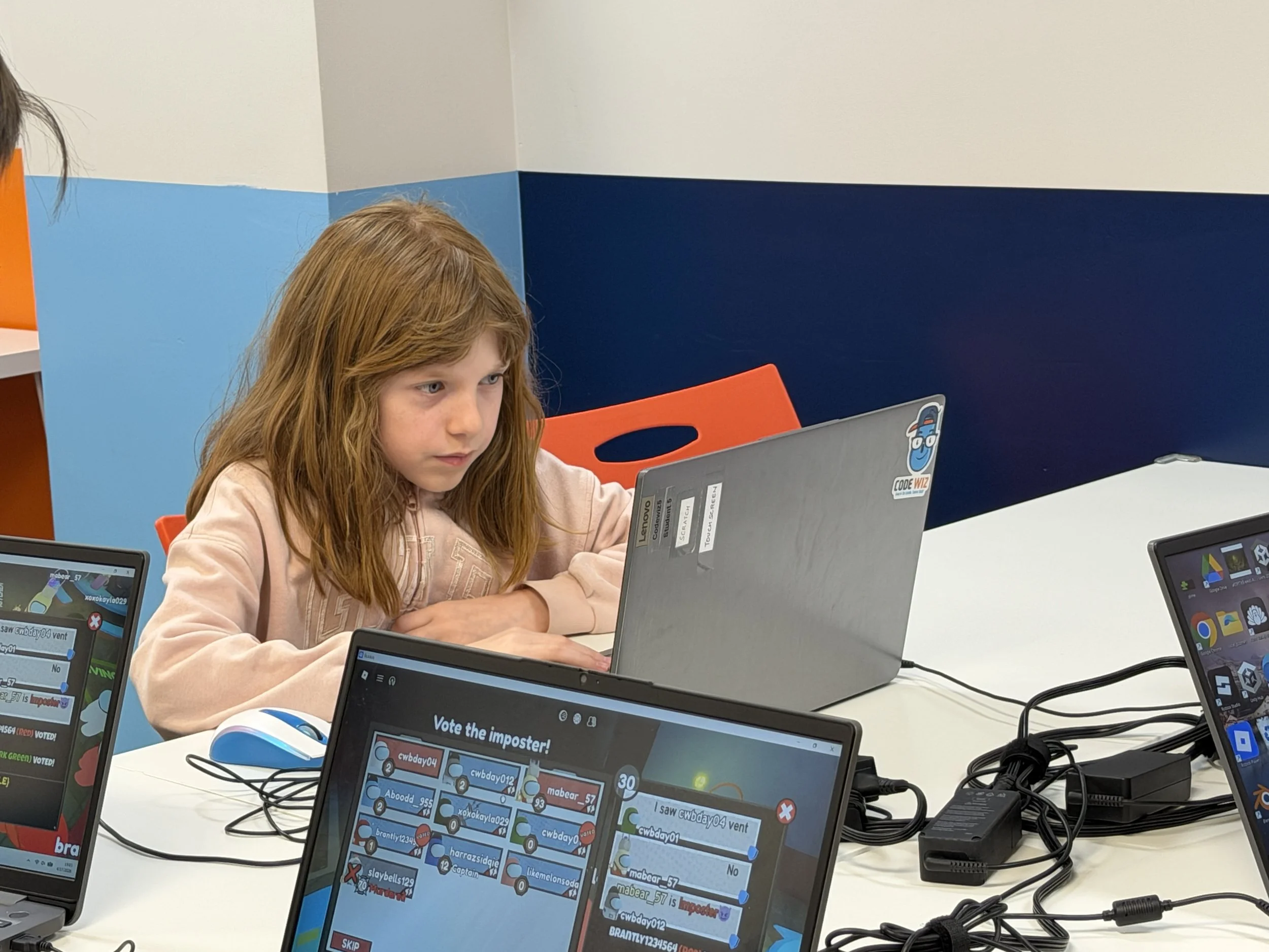 A young girl with red hair sitting at a white table, focused on a laptop with a sticker of a cartoon blue character with headphones. Multiple monitors display screens with game-like graphics and chat messages, connected by wires.