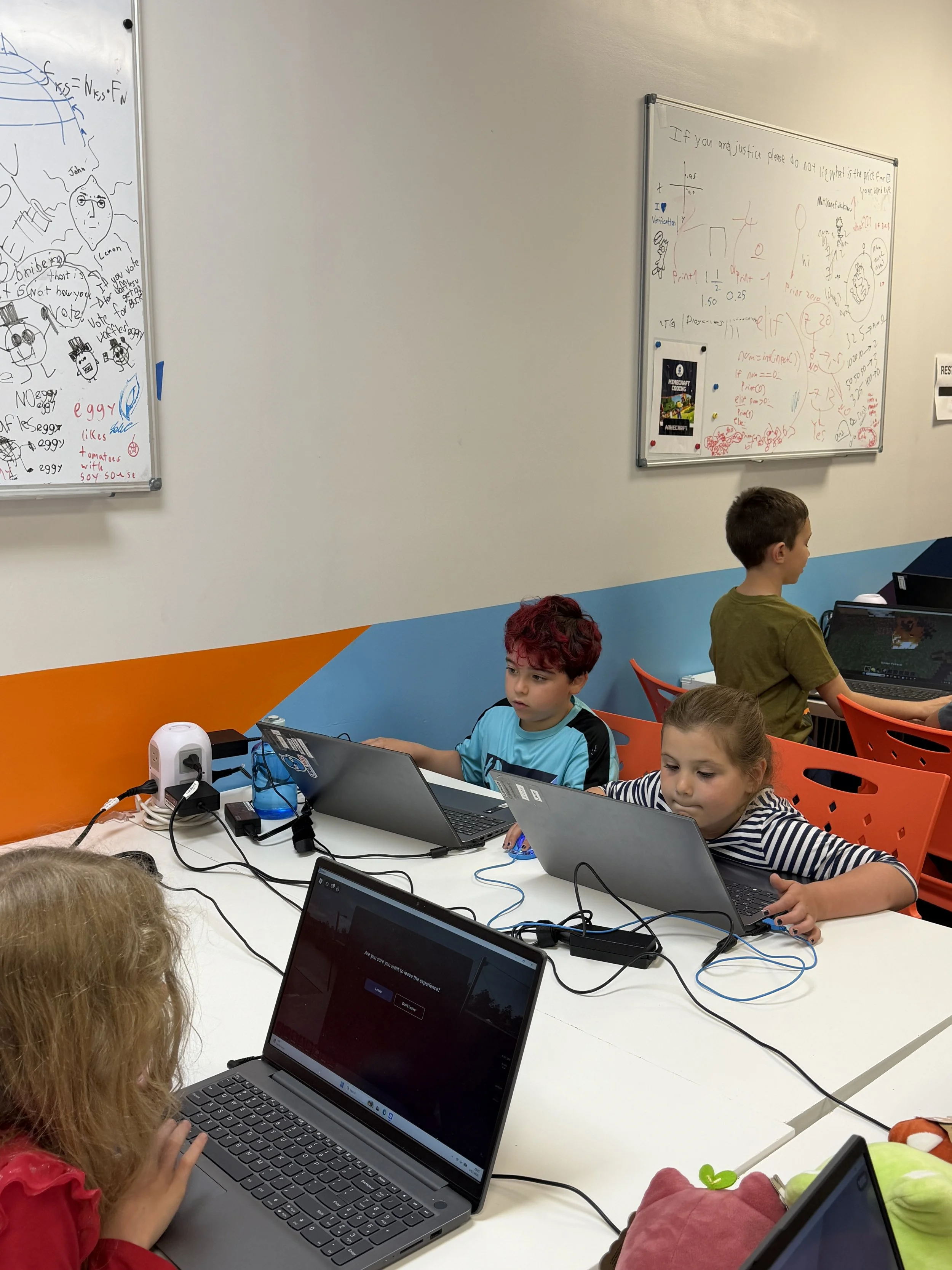 Young children sitting at a long white table working on laptops in a classroom with whiteboards and colorful walls.