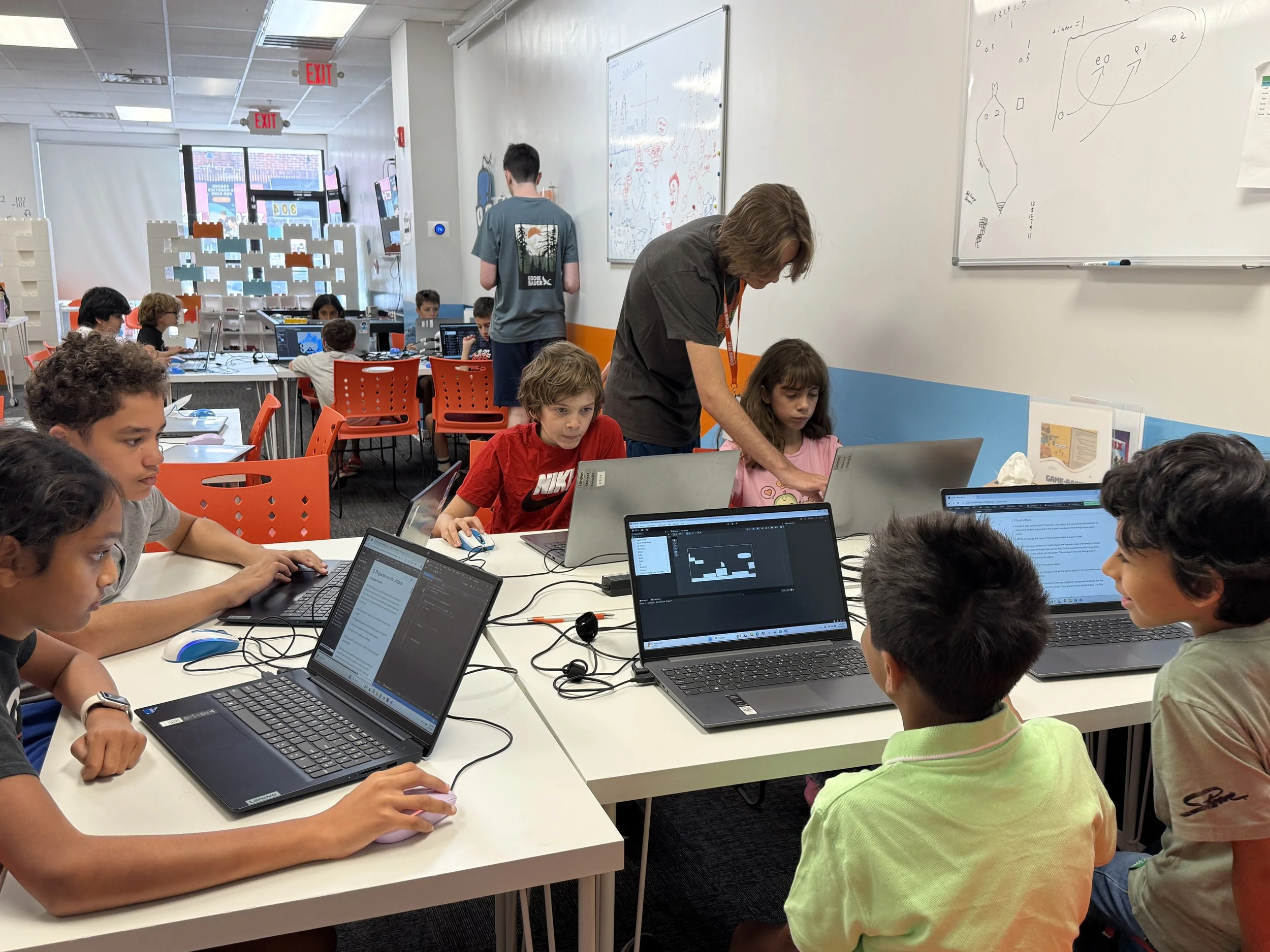 Children working on laptops in a classroom with math diagrams on whiteboards, a teacher assisting students, and others in the background using computers.
