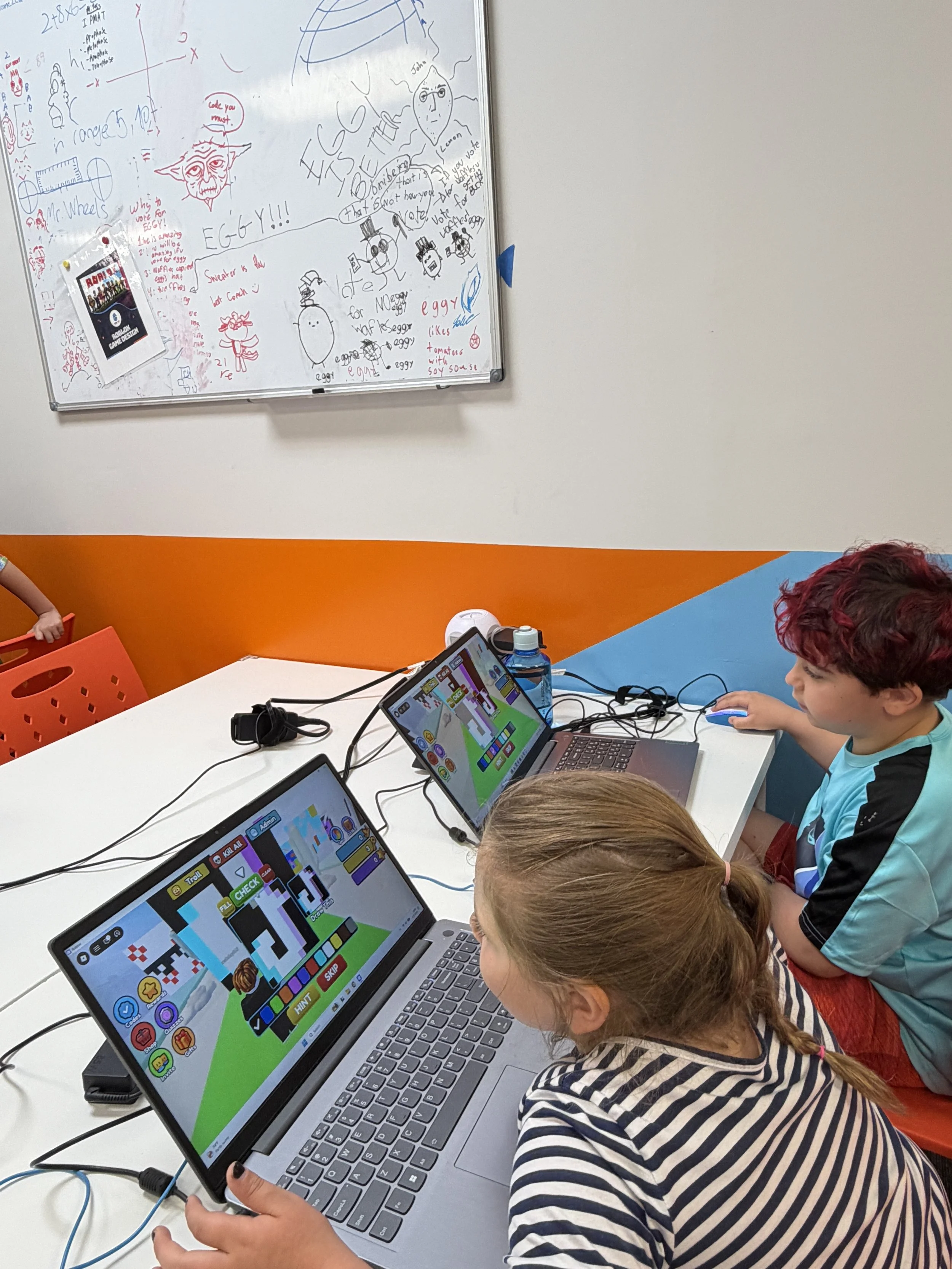Children working on laptops in a classroom, with a whiteboard filled with drawings and notes behind them.