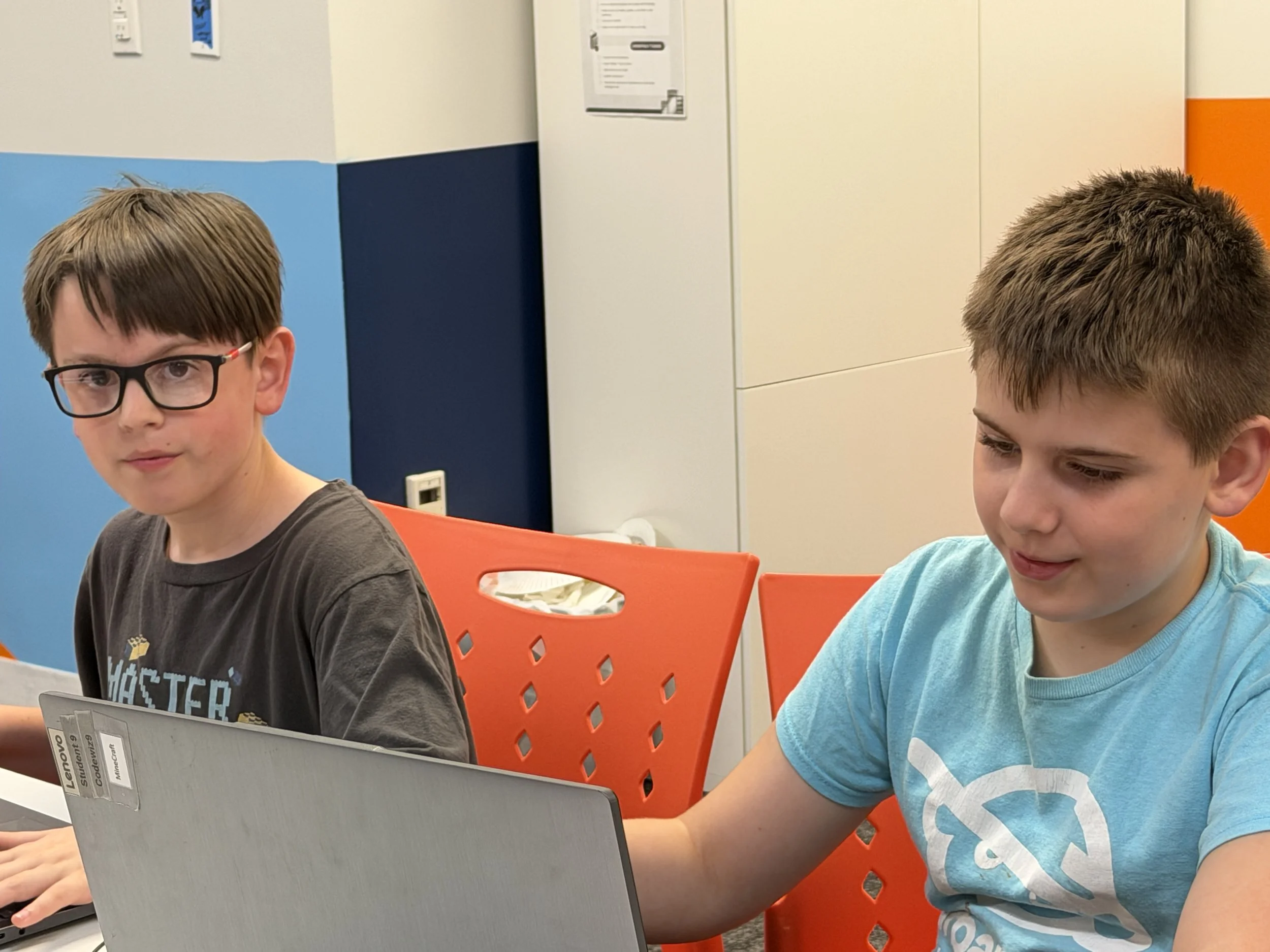 Two boys sitting at a table working on laptops in a colorful room with blue, white, and orange walls.