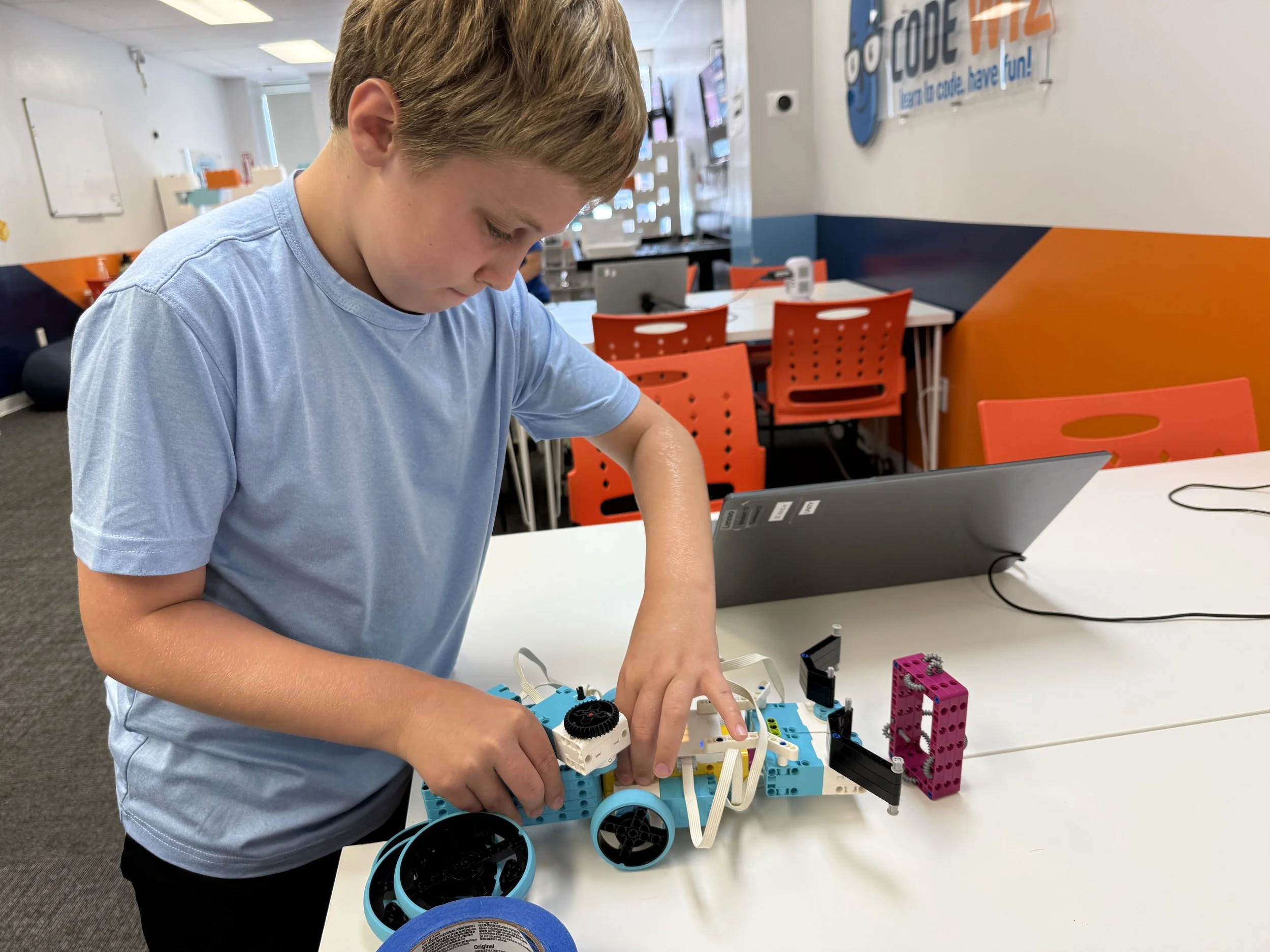 A young boy in a light blue shirt working on assembling a robot using LEGO bricks and components at a white table in a classroom or makerspace.