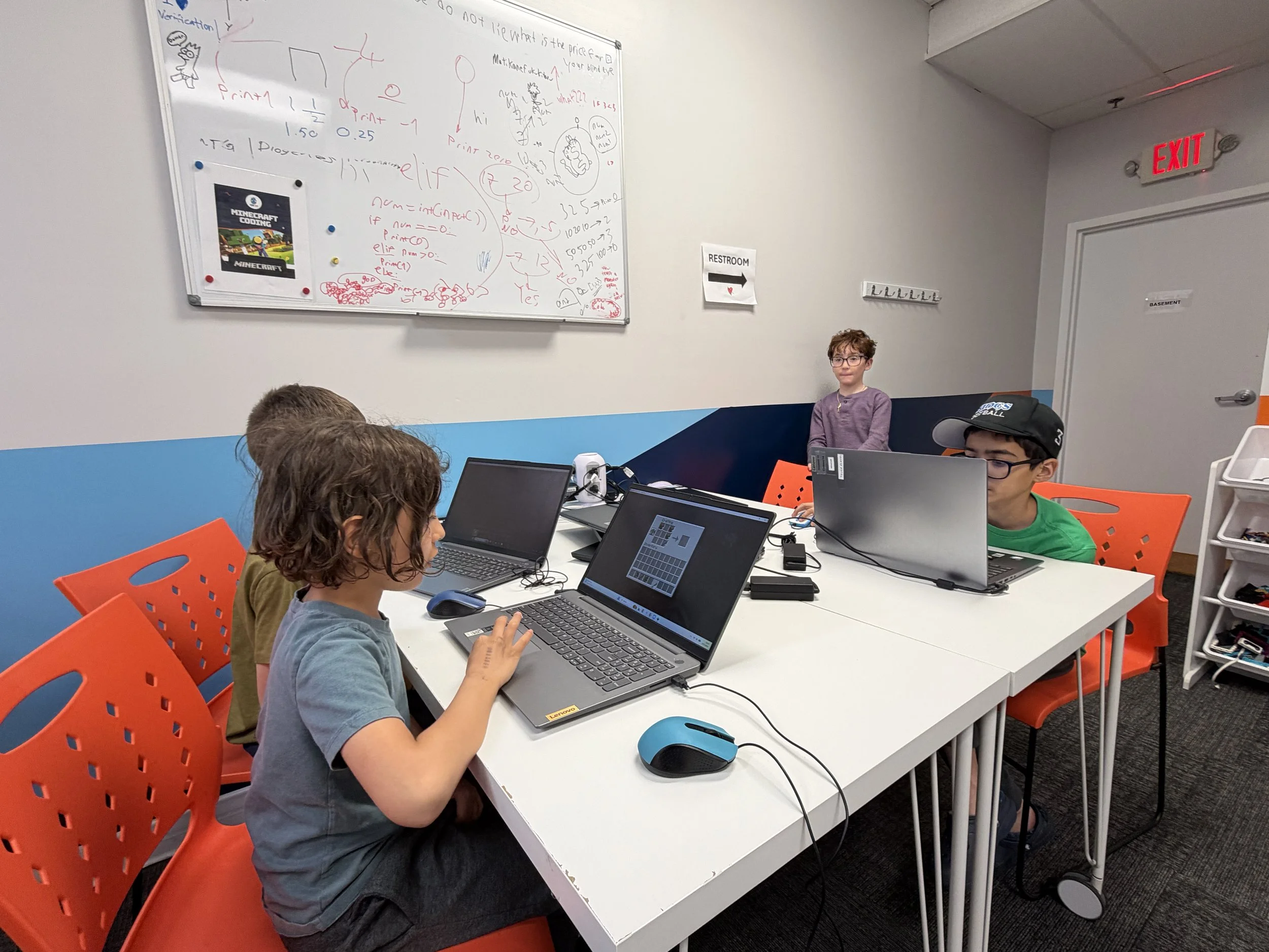 Group of children working on laptops in a classroom with a whiteboard filled with red and black drawings and notes, a sign pointing to restrooms, and orange chairs.