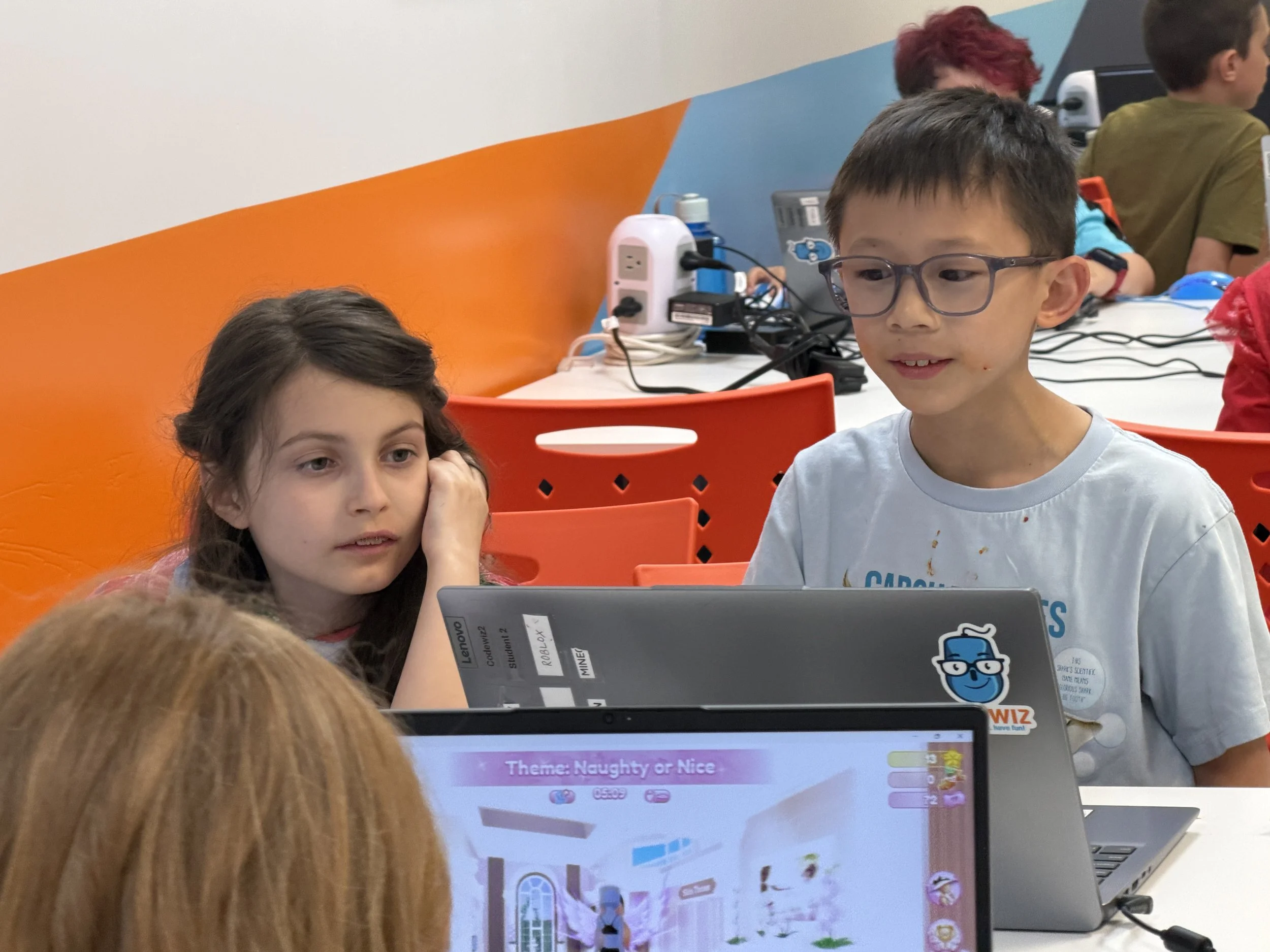 Two children, a girl with long brown hair and a boy with glasses, sit at a table with laptops in a classroom or computer lab. The girl is resting her head on her hand, and the boy is looking at the laptop screen. Other children and computer devices a