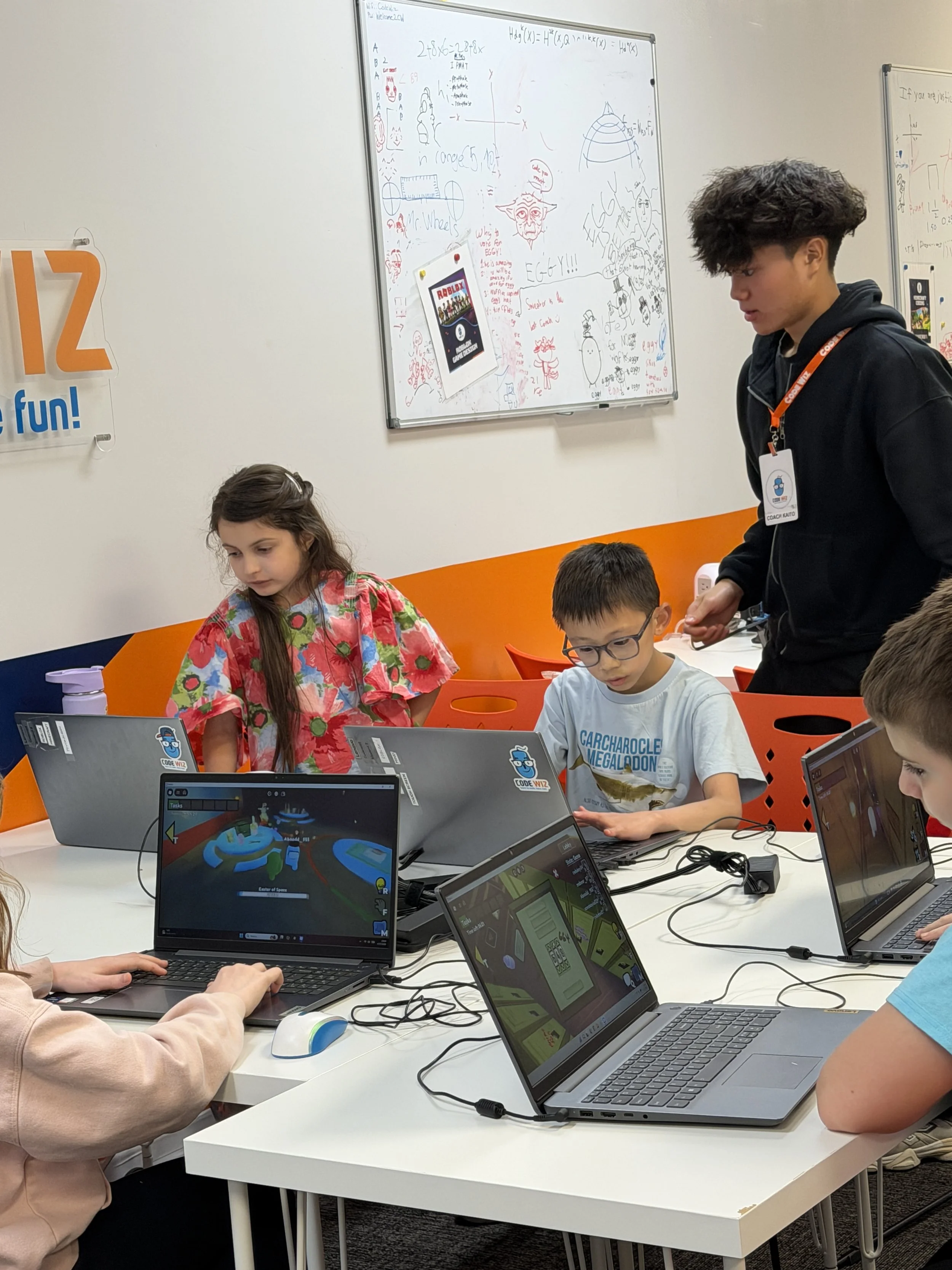 Children working on laptops in a classroom while a teacher supervises. The whiteboard behind them has various drawings and notes. The classroom has orange and white walls, and a poster on the wall says '12' and 'fun!'.