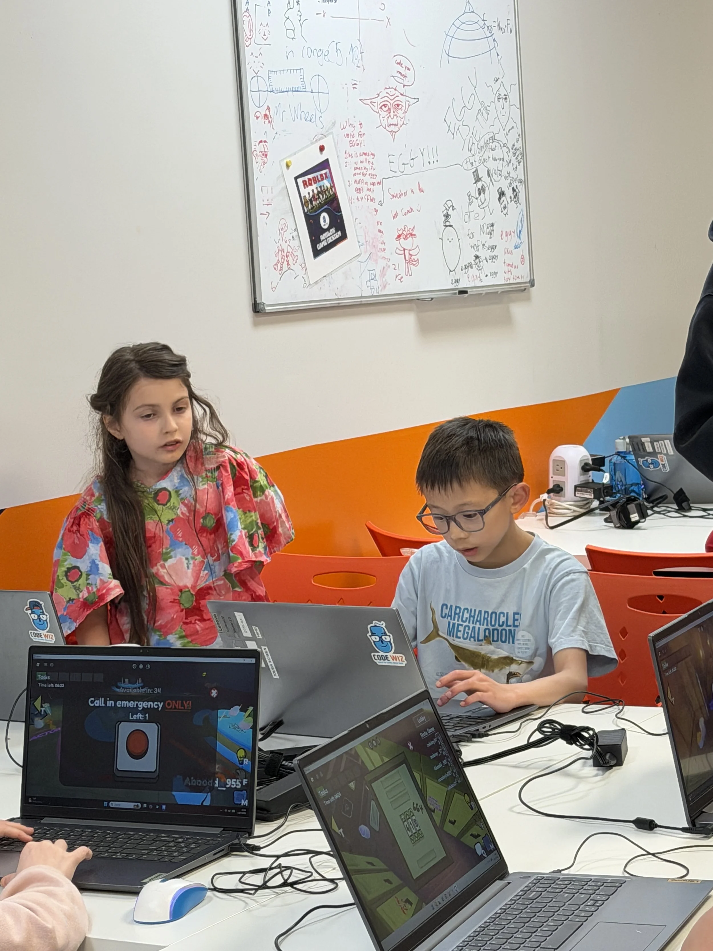 Two children working on laptops in a classroom, with a whiteboard in the background covered in sketches and doodles.