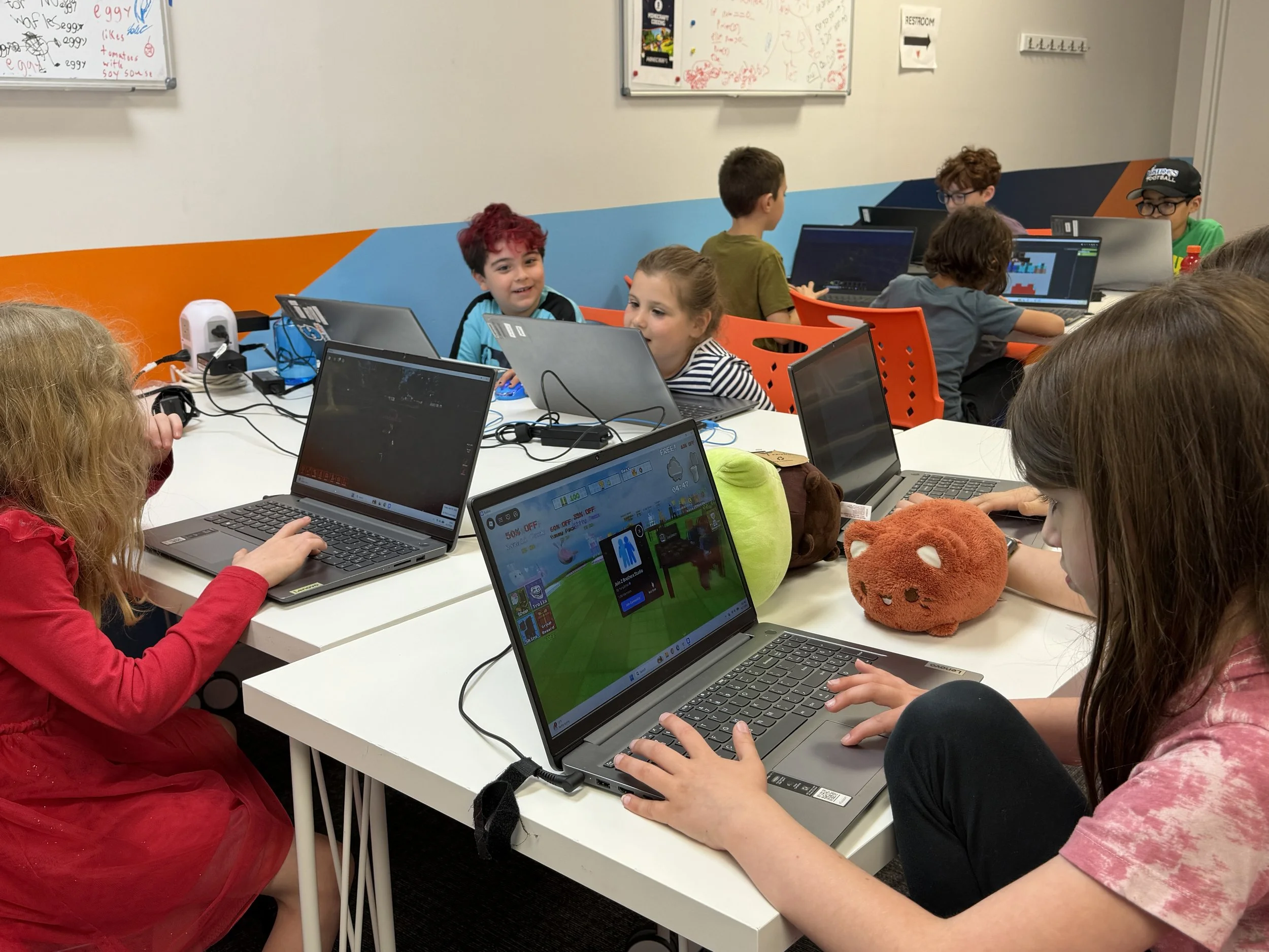 A group of children sitting at a table using laptops in a classroom with colorful walls.