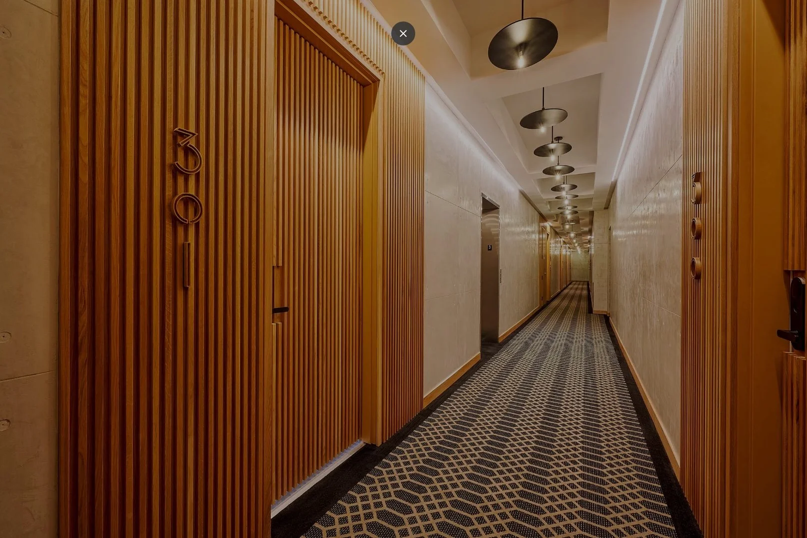 Clean hotel corridor with wooden wall paneling, elevator, patterned carpet, and modern ceiling lighting.