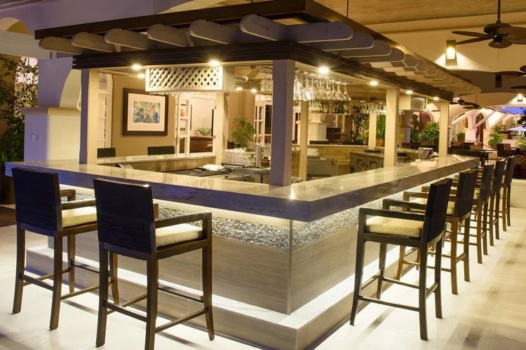 A modern indoor bar with a square counter, bar stools, hanging wine glasses, and backlit decor in a restaurant setting.