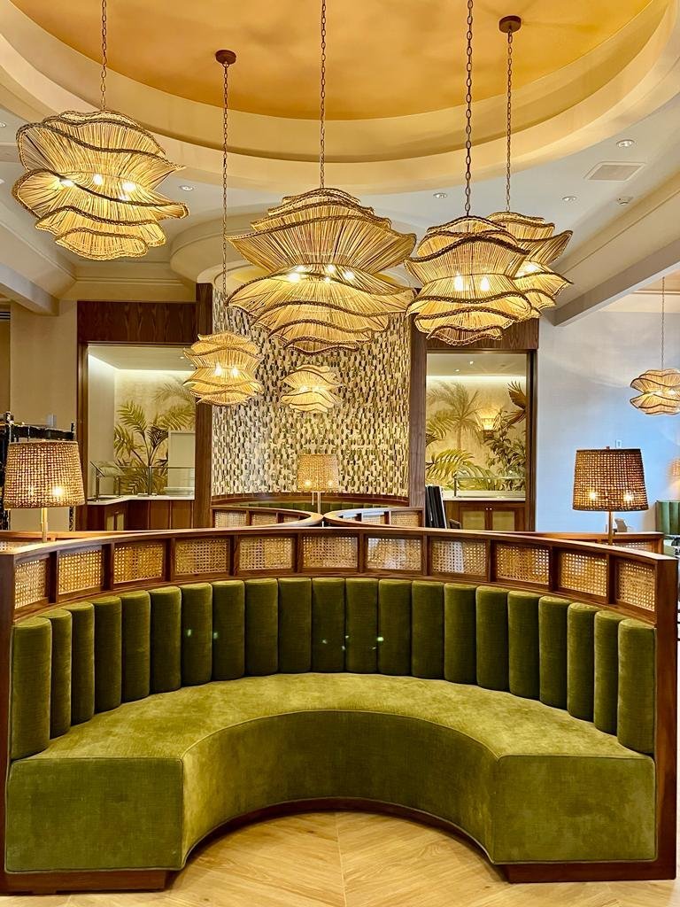 Elegant hotel lobby seating area with a curved green velvet sofa, decorative hanging pendant lights, and tropical-themed wall decor with plants.