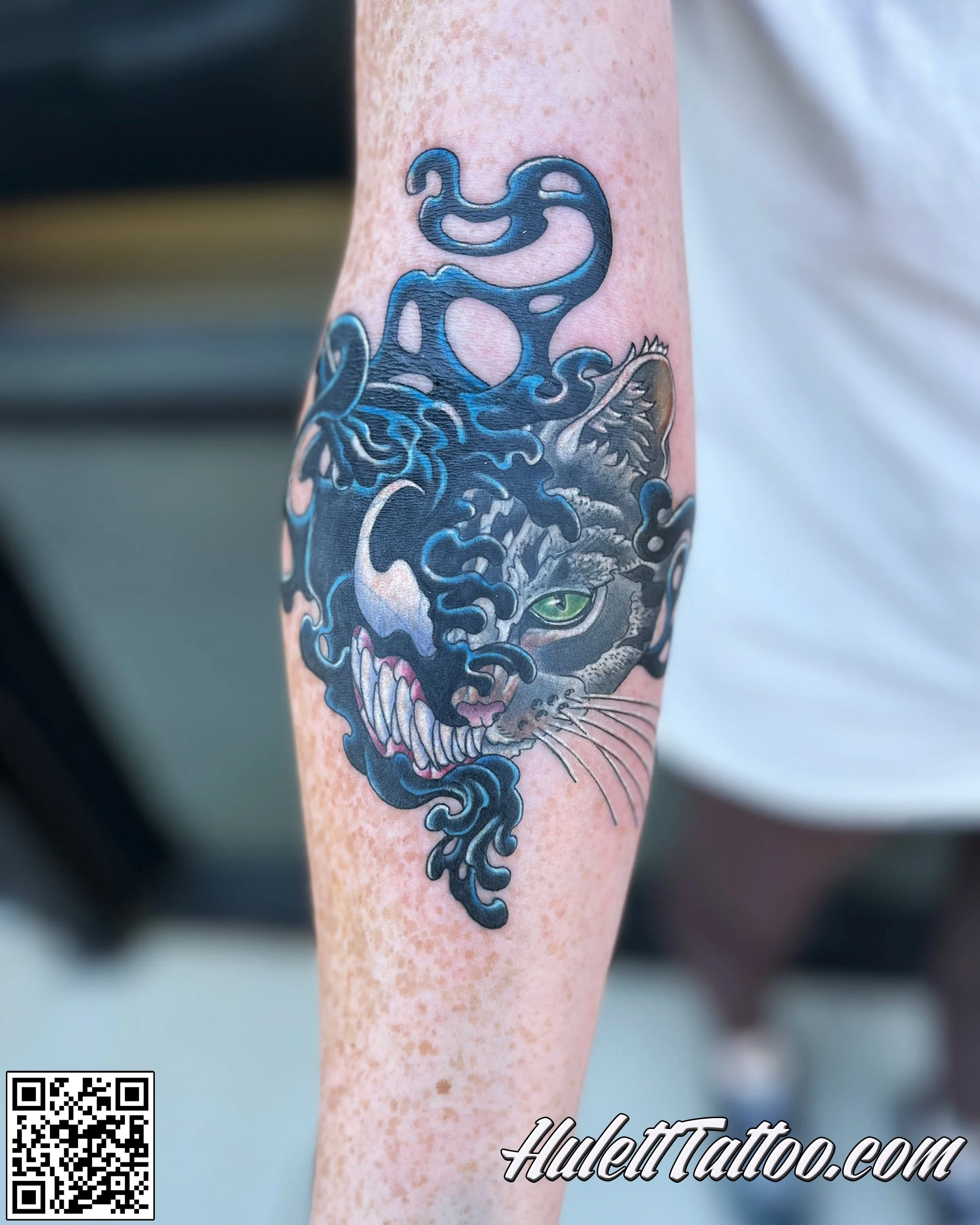 Venom Kitty tattoo of a snarling tiger's face with green eye, overlaid with black and blue smoke or ink swirling around and through it