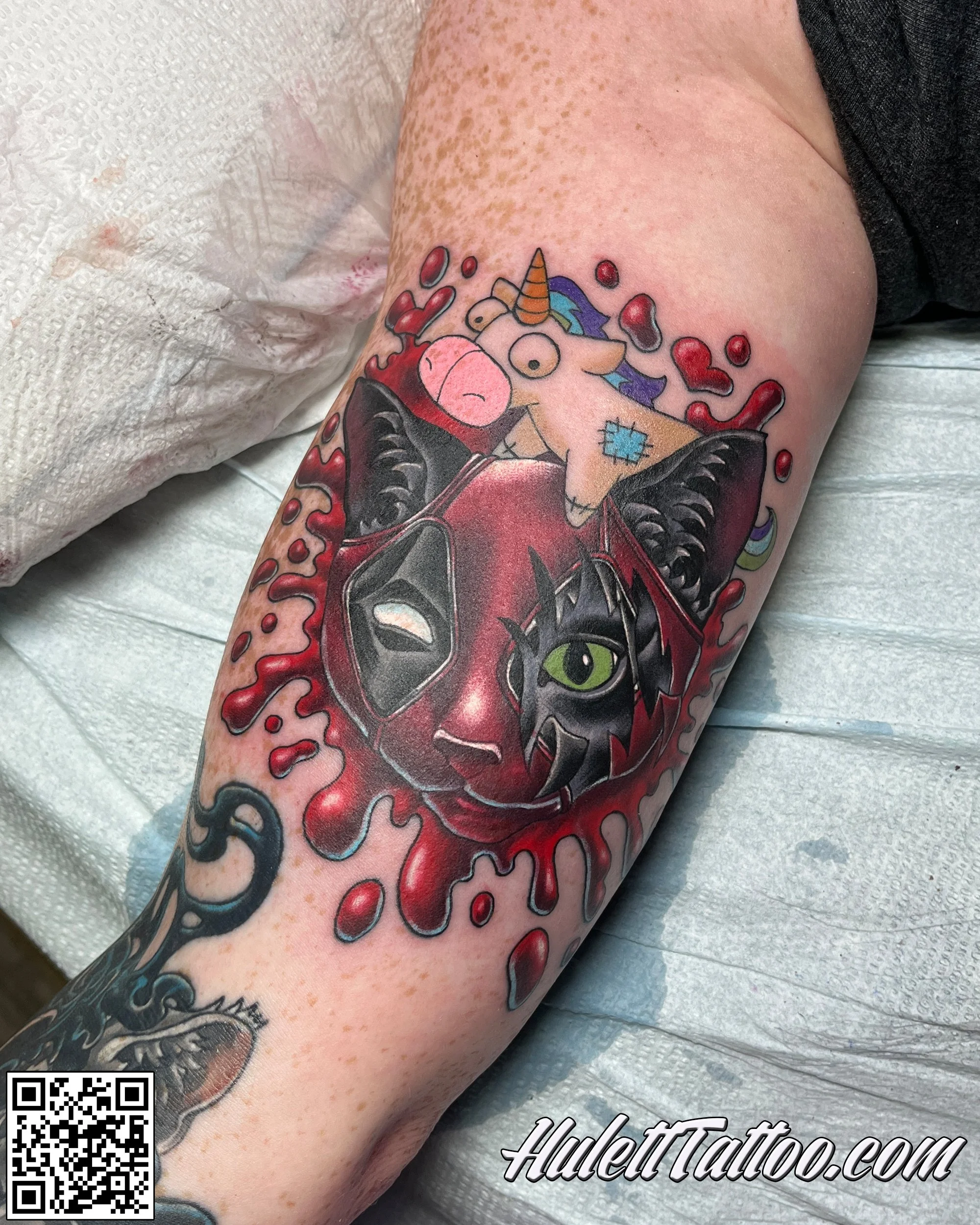 A colorful tattoo on a person's arm featuring a cat portrait done as DeadPool and cartoon unicorn with a rainbow mane, surrounded by red hearts, a black cat with green eyes, and the text 'HulettTattoo.com' and a QR code.