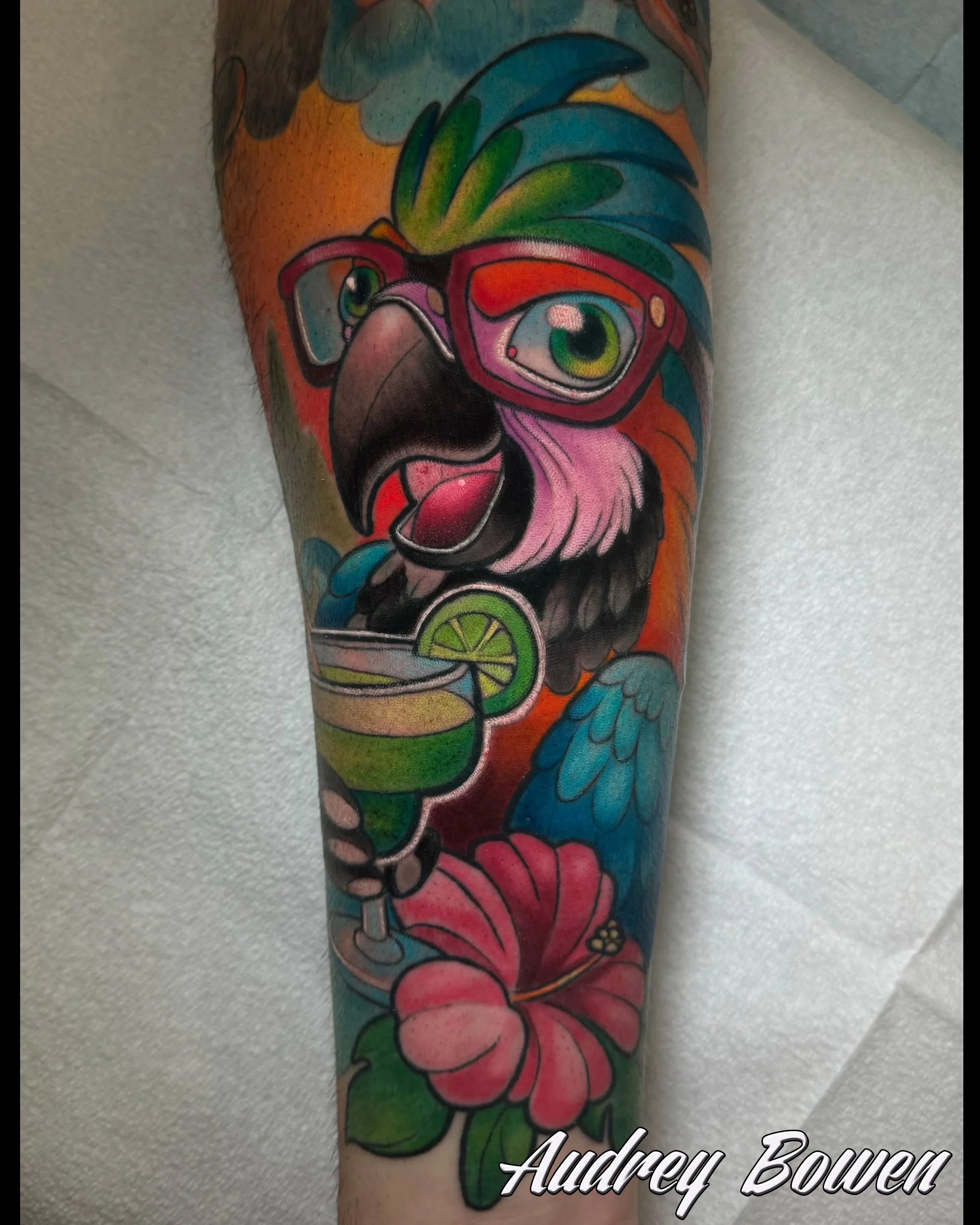Colorful tattoo of a parrot with glasses, surrounded by vibrant flowers and leaves. Tattoo by Audrey Bowen, Karmic Tattoo, Clearwater Florida