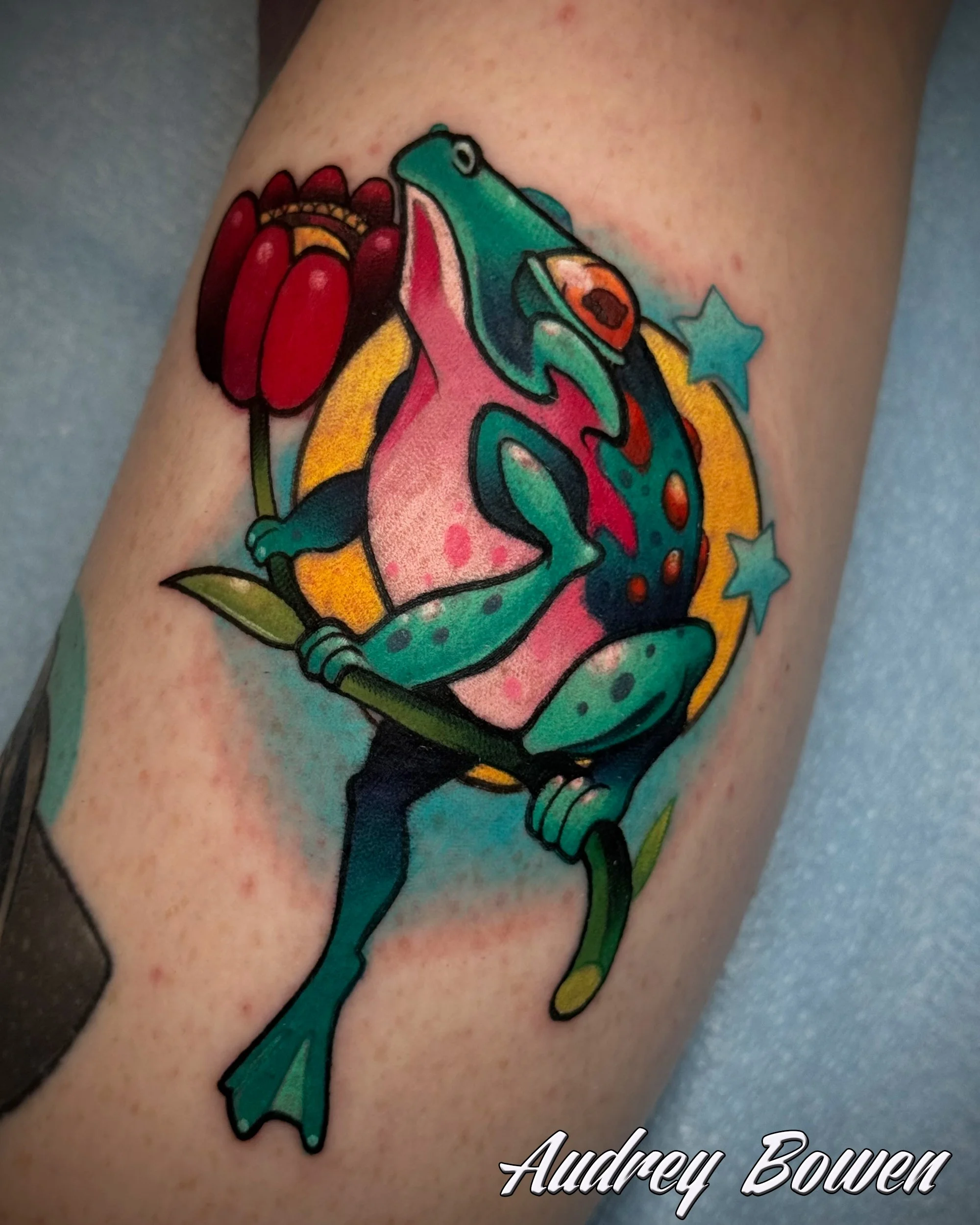 Colorful tattoo of a frog on skin, with stars and a flower design. Tattoo by Audrey Bowen, Karmic Tattoo, Clearwater Florida