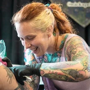 Audrey wearing her black gloves, holding a tattoo machine and working on a person's arm in a tattoo studio.