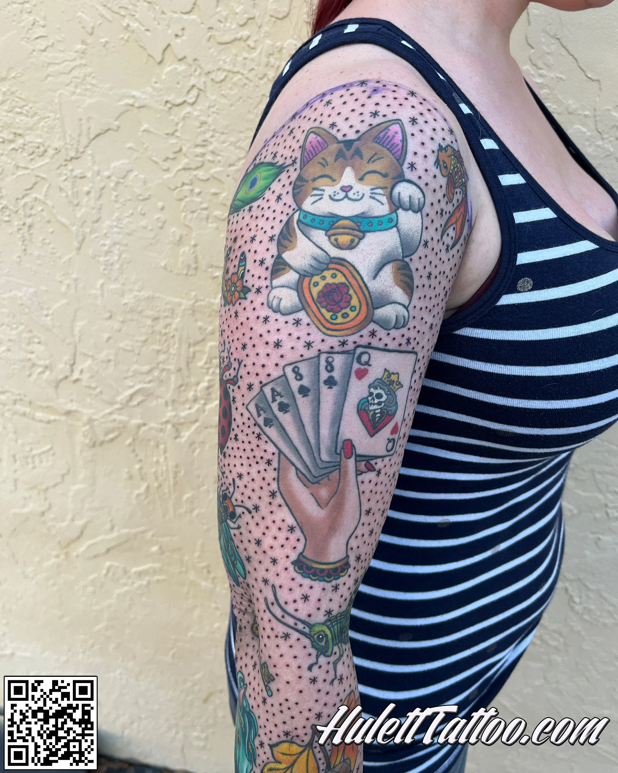 Colorful shoulder tattoo featuring a Maneki-neko (lucky cat), playing cards, and various small decorative elements on a person's arm done in a style of traditional tattoo.
