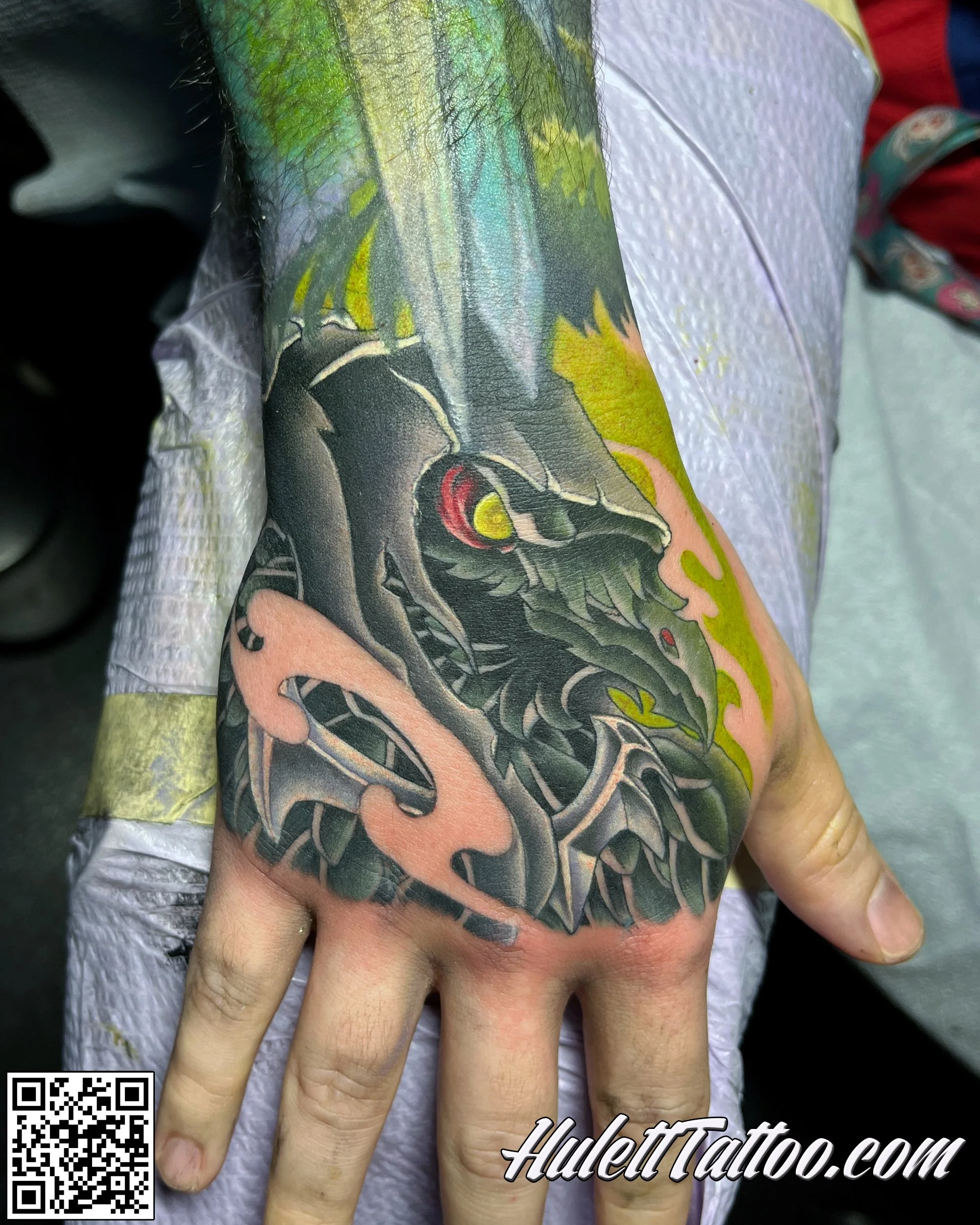 A close-up of a person's arm with a colorful tattoo of a dragon. The person's hand is resting on their forehead, and they are wearing a white shirt. The background shows part of a red and blue garment and a white textured surface.
