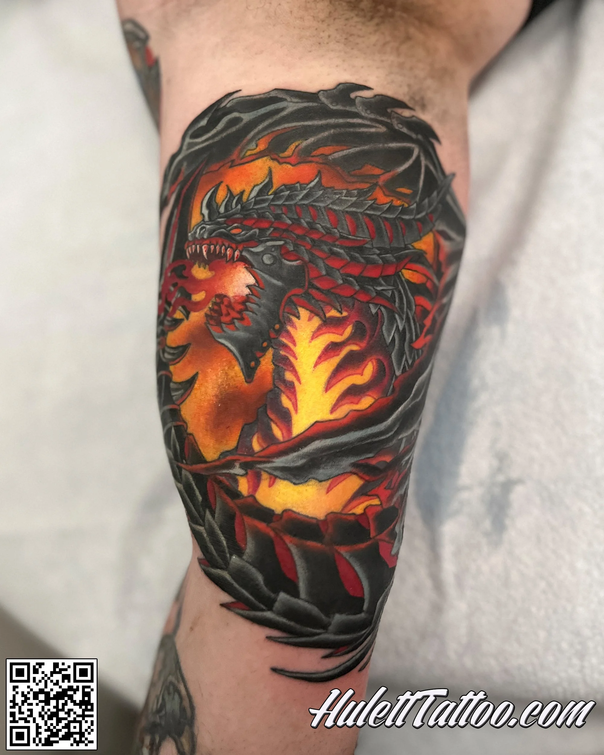 Tattoo of a fierce black and red dragon with sharp teeth, on an orange and yellow fiery background, around a person's arm.