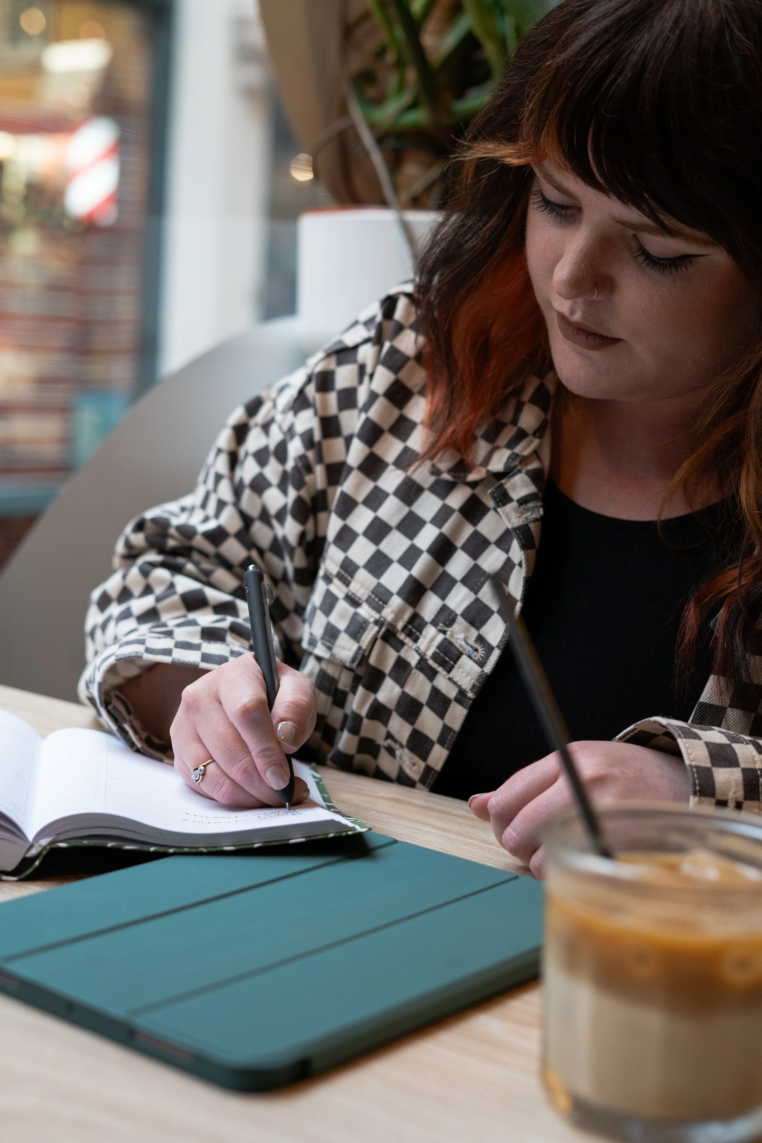 Bloom Creative Founder Courtney Farrow writing in a note book. Her hair is brown and orange, she's wearing a black and white checkerboard jacket and on the table in front of her is a note pad, green iPad and iced oat latte.