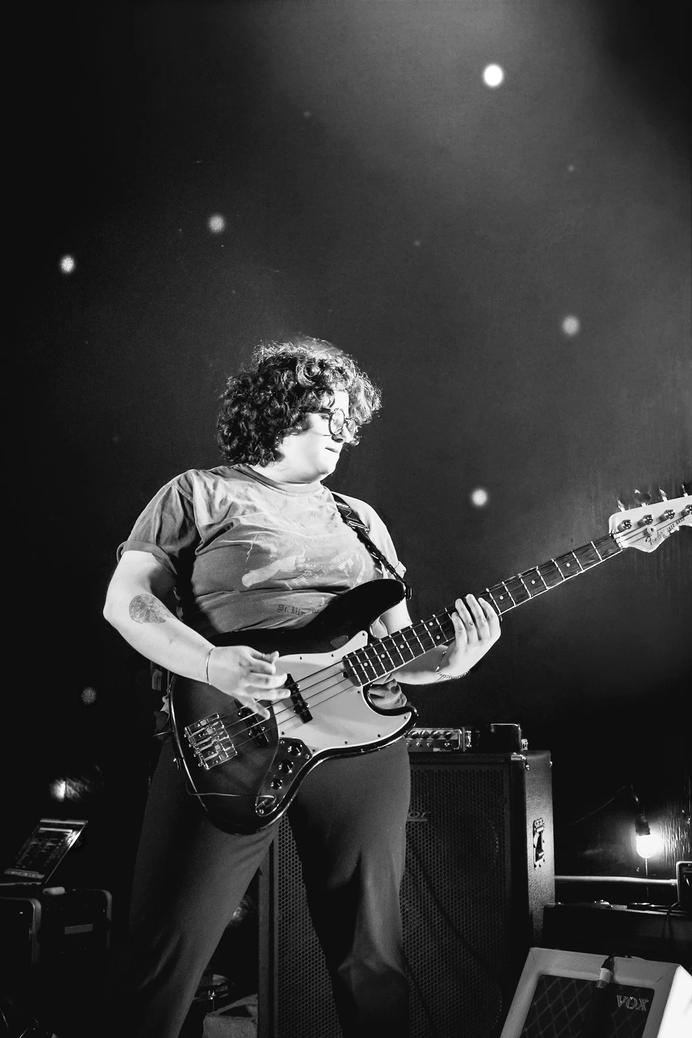 A woman with curly hair and glasses playing an electric bass guitar on stage, with amplifier equipment in the background.