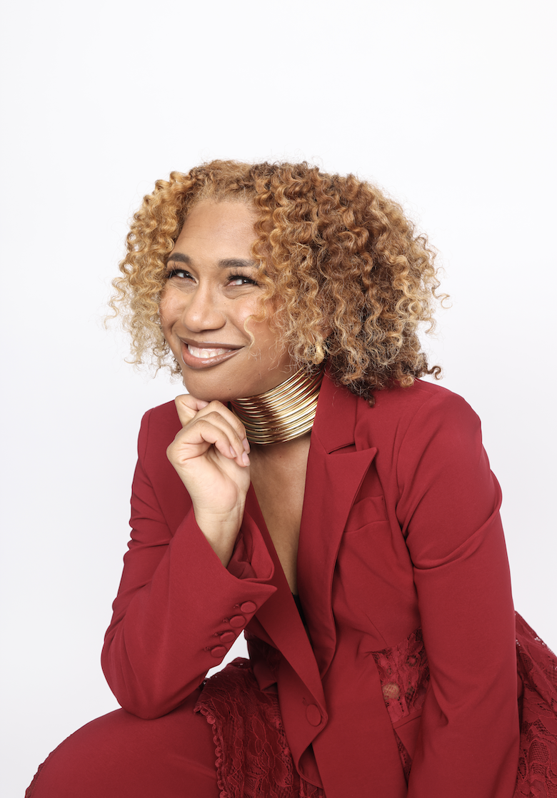 A woman with curly blonde hair, wearing a red blazer and matching red dress with lace details, smiling and posing against a white background.