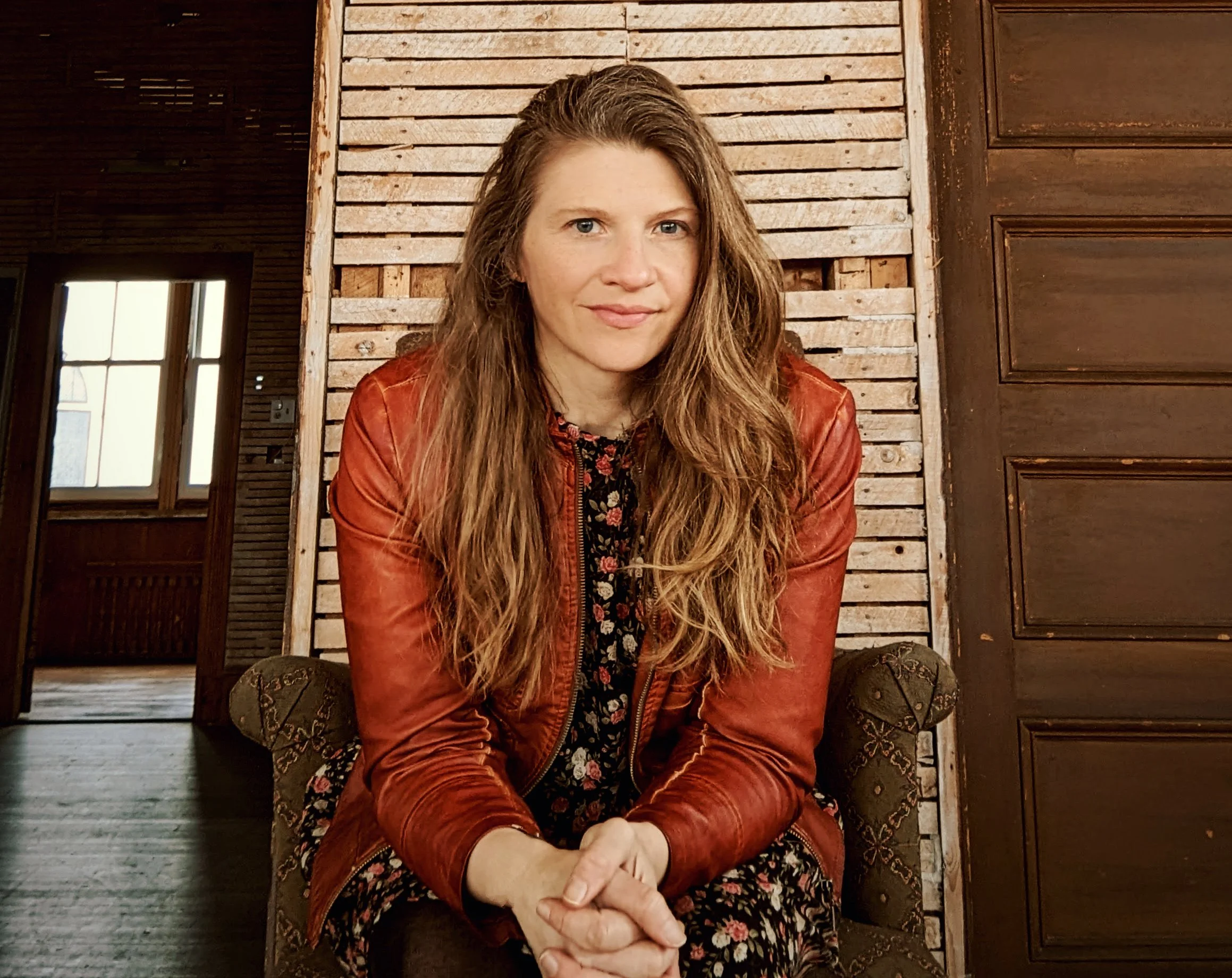 A woman with long wavy hair, wearing a red leather jacket, sitting on a patterned chair in a rustic room with wooden walls and natural light coming through the window.