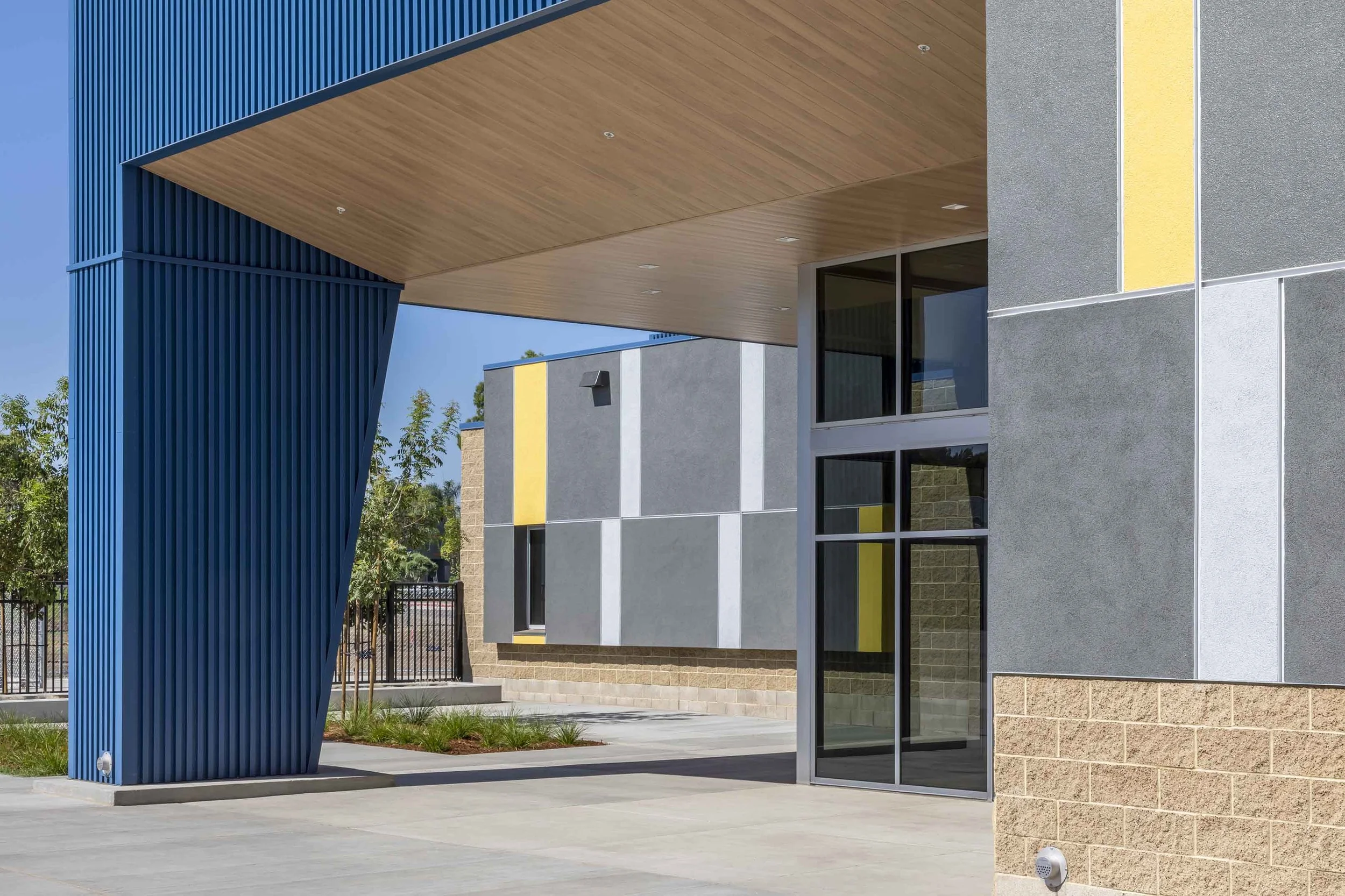 Modern architectural building with blue vertical metal beams, wooden ceiling overhang, gray walls with yellow accents, and large glass doors.