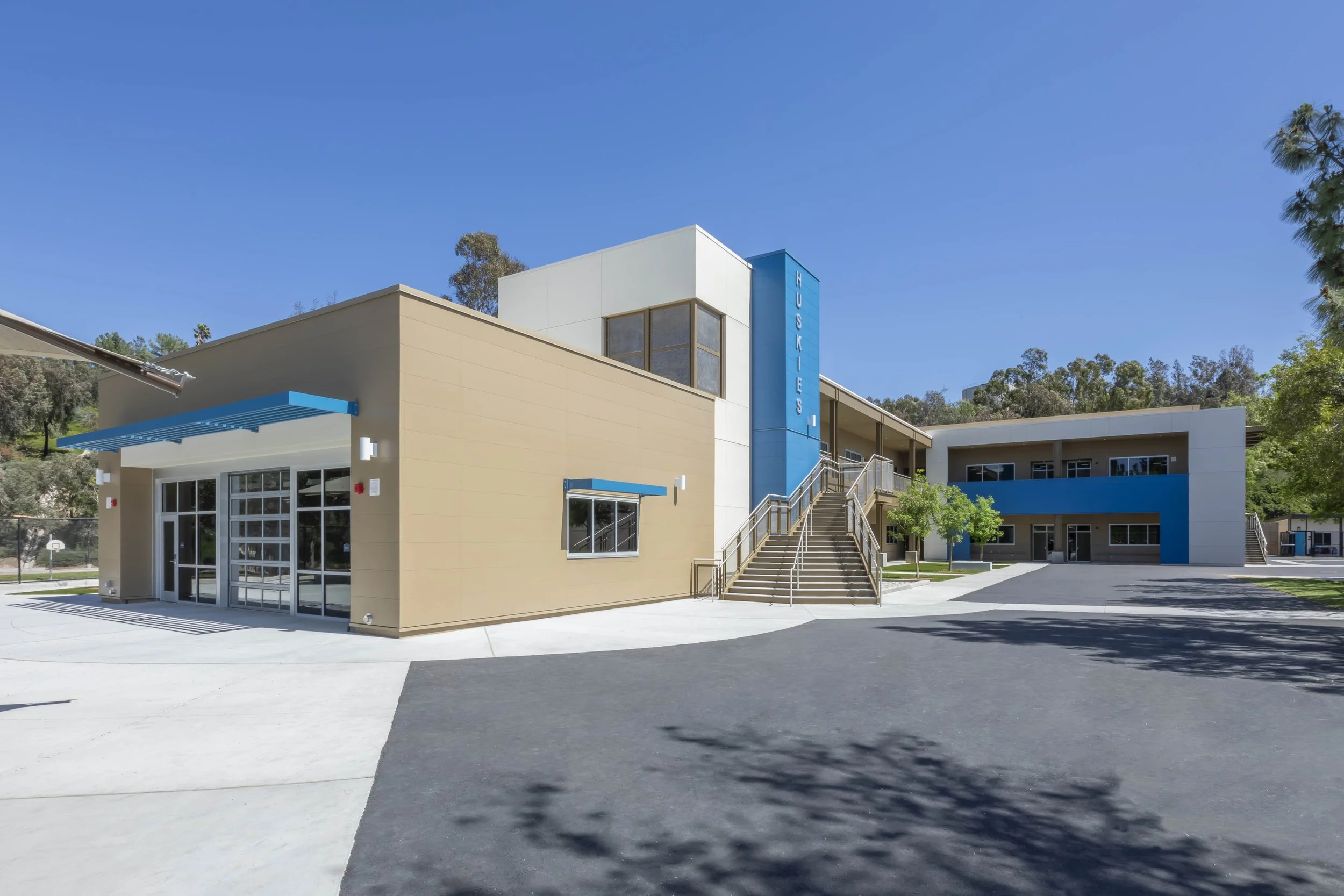 Modern school building with beige and white exterior, blue accents, glass doors, and stairs leading to classrooms, with a basketball court visible in the background.