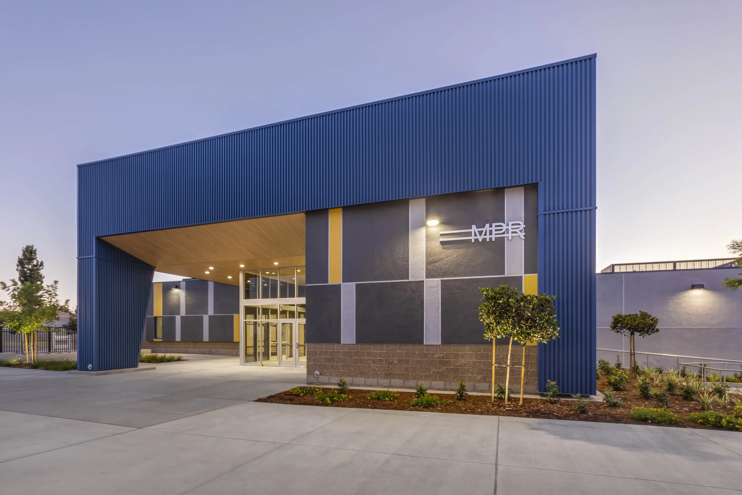 Modern building with blue and gray exterior, trees, and pathway at dusk, with illuminated sign that reads 'MPA'